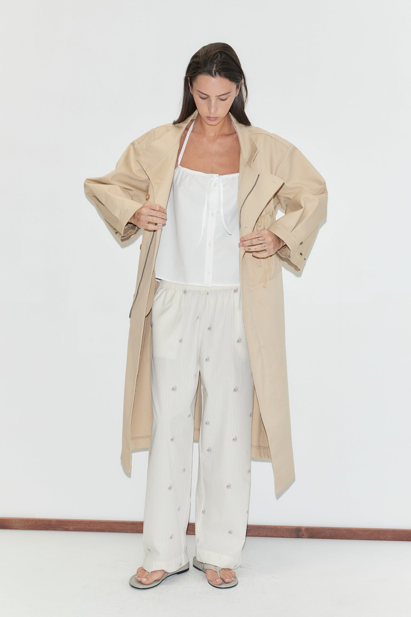 A woman stands against a white wall, wearing a beige trench coat over a white top and the Ease Trouser - Floral Stripe by Deiji Studios. She is looking down and wearing flip-flops.