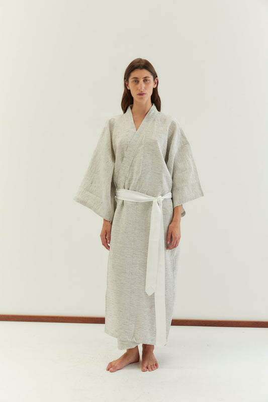 A person stands barefoot against a plain white background, wearing the 02 robe - pinstripe by Deiji Studios, featuring wide sleeves and a soft linen waist tie.