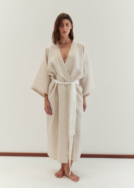 A woman stands barefoot against a plain white background, wearing the 02 robe - oatmeal by Deiji Studios. This loose, light beige robe features wide sleeves and a linen waist tie. She faces the camera with a neutral expression.