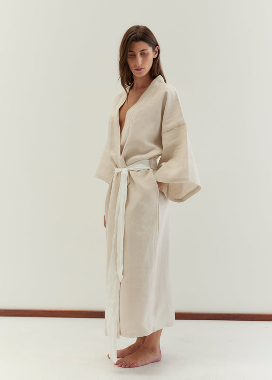 A woman stands barefoot on a light floor, wearing Deiji Studios' the 02 robe - oatmeal, a full-length linen robe with wide sleeves and a white fabric belt, set against a plain white background.