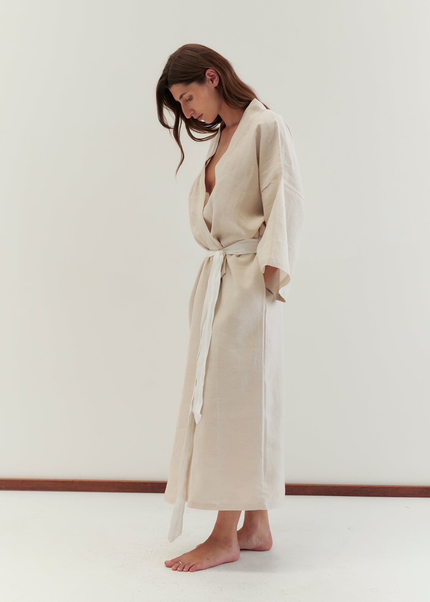 A woman with long brown hair stands barefoot against a plain white background, wearing the 02 robe - oatmeal by Deiji Studios, featuring wide sleeves and a linen waist tie, gazing downward in a relaxed pose.
