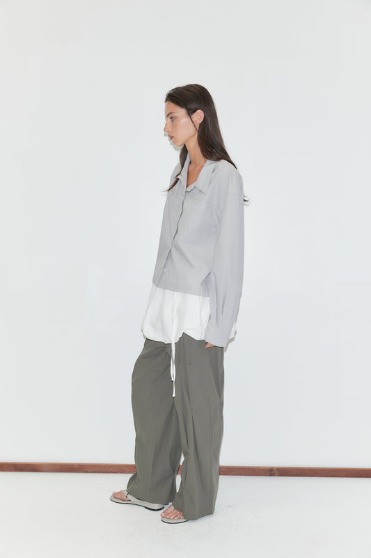 A woman with long dark hair stands in profile against a plain white background wearing the Boxy Shirt - Cloud Stripe by Deiji Studios, a pale grey-blue fine-striped boxy shirt, over a white top with olive wide-leg trousers and grey flip-flops.