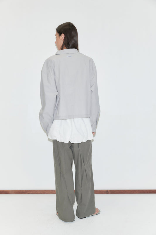 A woman with long dark hair stands with her back to the camera against a plain white background wearing the Boxy Shirt - Cloud Stripe by Deiji Studios, a pale grey-blue fine-striped boxy shirt, over a white top with olive wide-leg trousers.