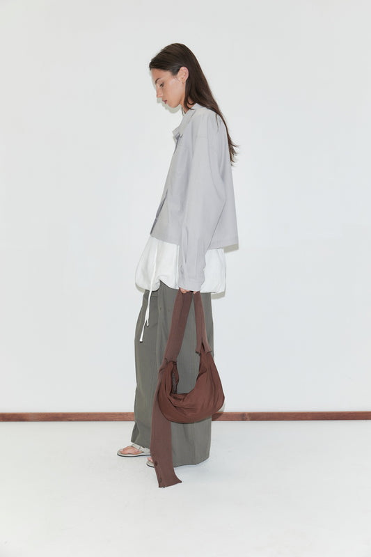 A woman with long dark hair stands in profile against a plain white background wearing the Boxy Shirt - Cloud Stripe by Deiji Studios over a white top with olive wide-leg trousers, carrying a brown shoulder bag.