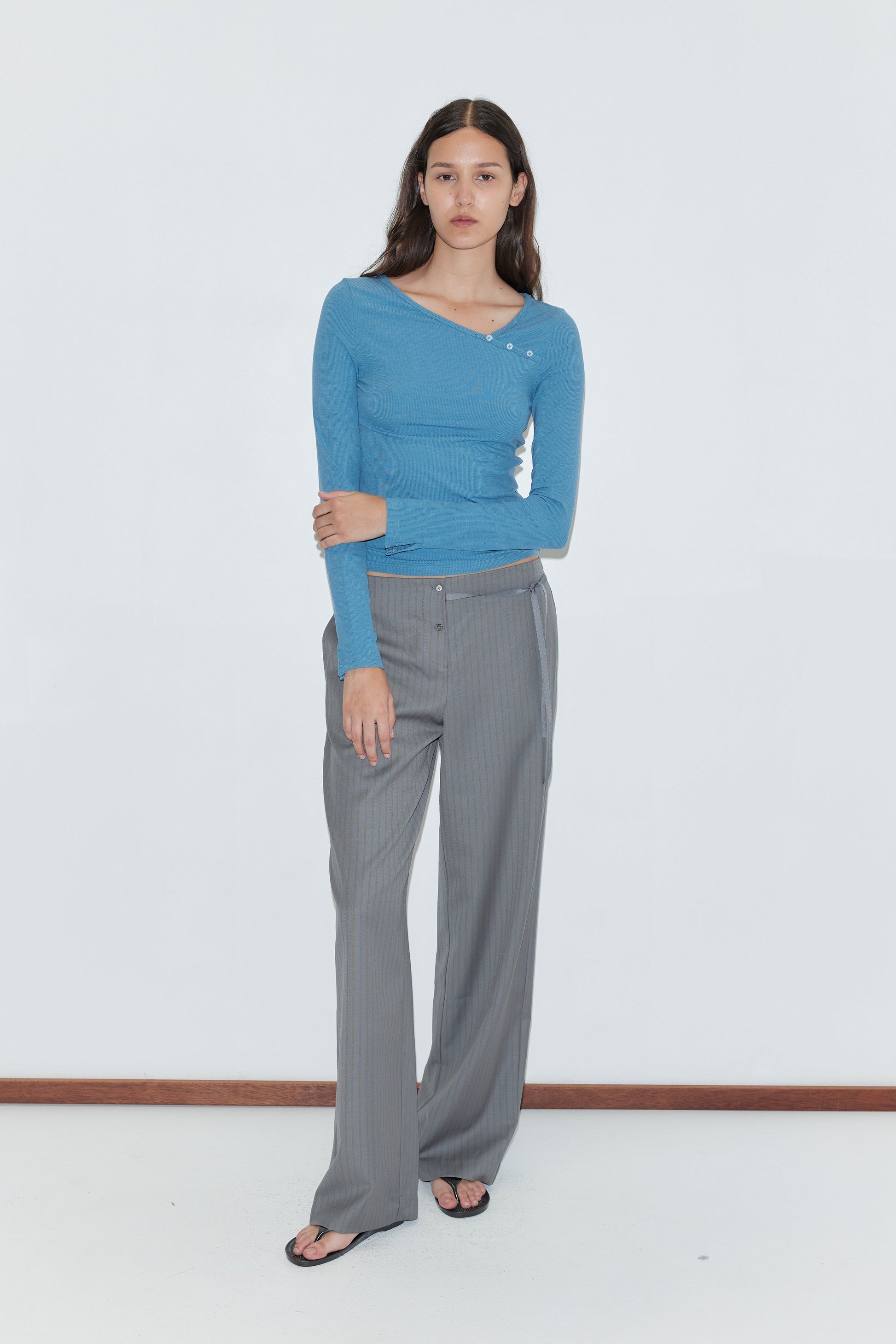 A woman with long dark hair stands against a white background, wearing the Deiji Studios Multi Button Top in Aqua with gray wide-leg trousers and black flip-flops. The top features long sleeves and an asymmetric neckline.