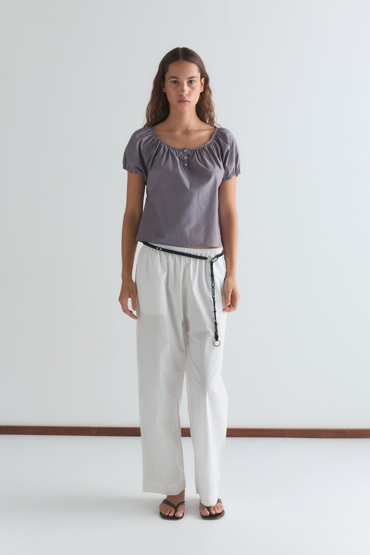 A woman with long, wavy brown hair and a neutral expression stands against a white background wearing the Poplin T Shirt - Currant by Deiji Studios, paired with loose white pants, a black belt, and black sandals.