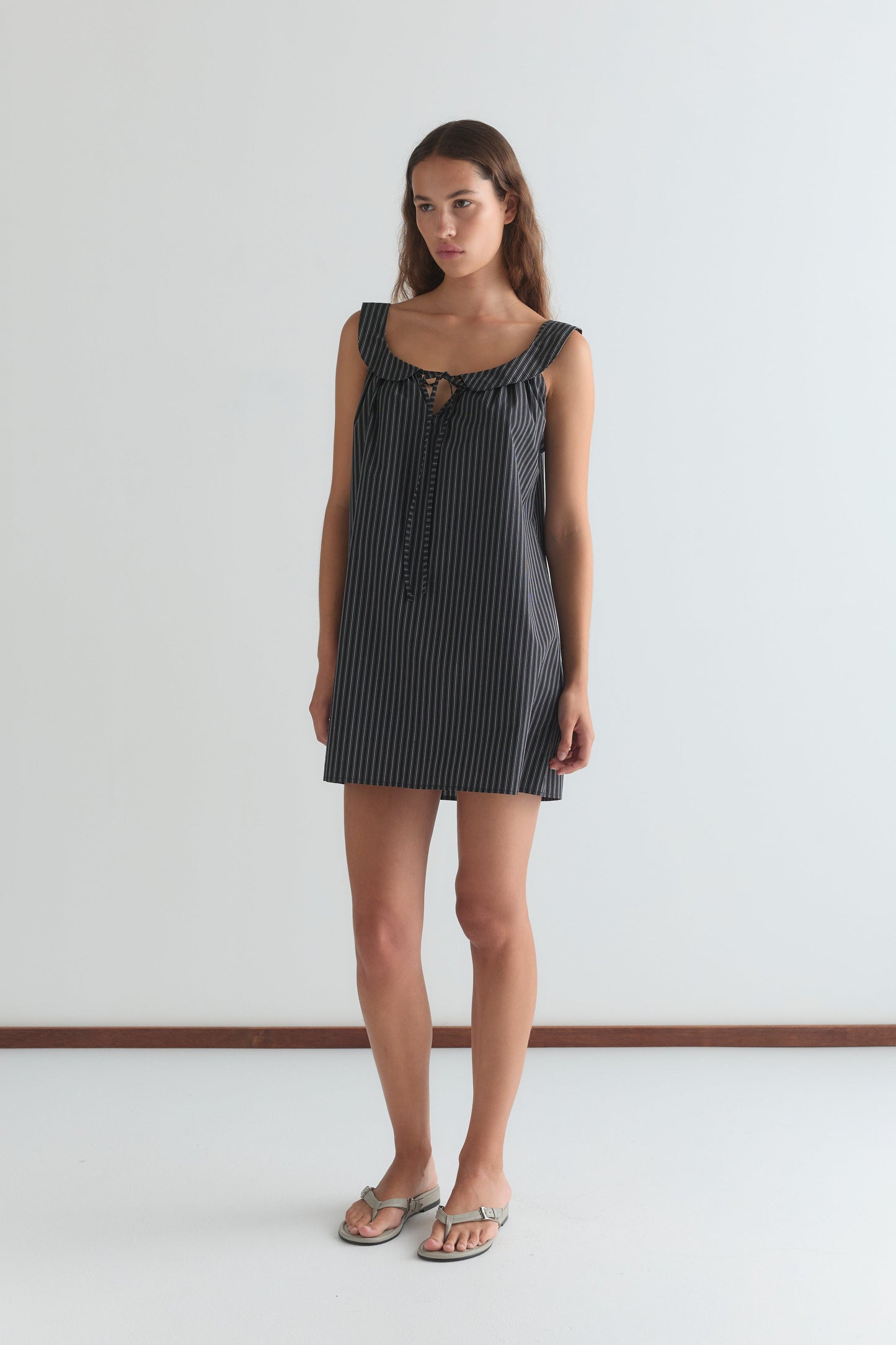 A woman stands against a white wall wearing the Rounded Neck Dress - Mono stripe by Deiji Studios, featuring a sleeveless design, rounded neck, front tie detail, and mono stripe pattern. She has long hair, a neutral expression, and wears sandals.