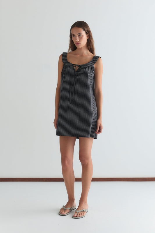 A woman stands against a white wall wearing the Rounded Neck Dress - Mono stripe by Deiji Studios, featuring a sleeveless design, rounded neck, front tie detail, and mono stripe pattern. She has long hair, a neutral expression, and wears sandals.