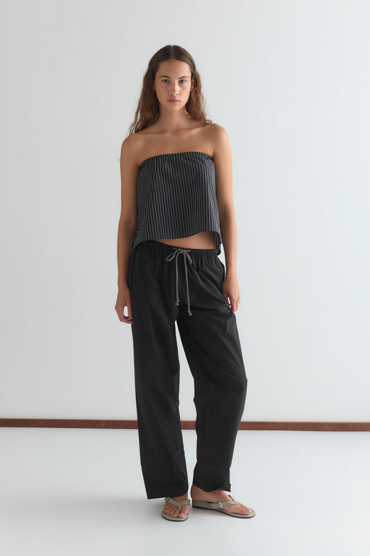 A woman with long, wavy brown hair and a neutral expression stands against a plain white wall wearing a black and white strapless top, the Layered Pant - Black by Deiji Studios, and flip-flops.