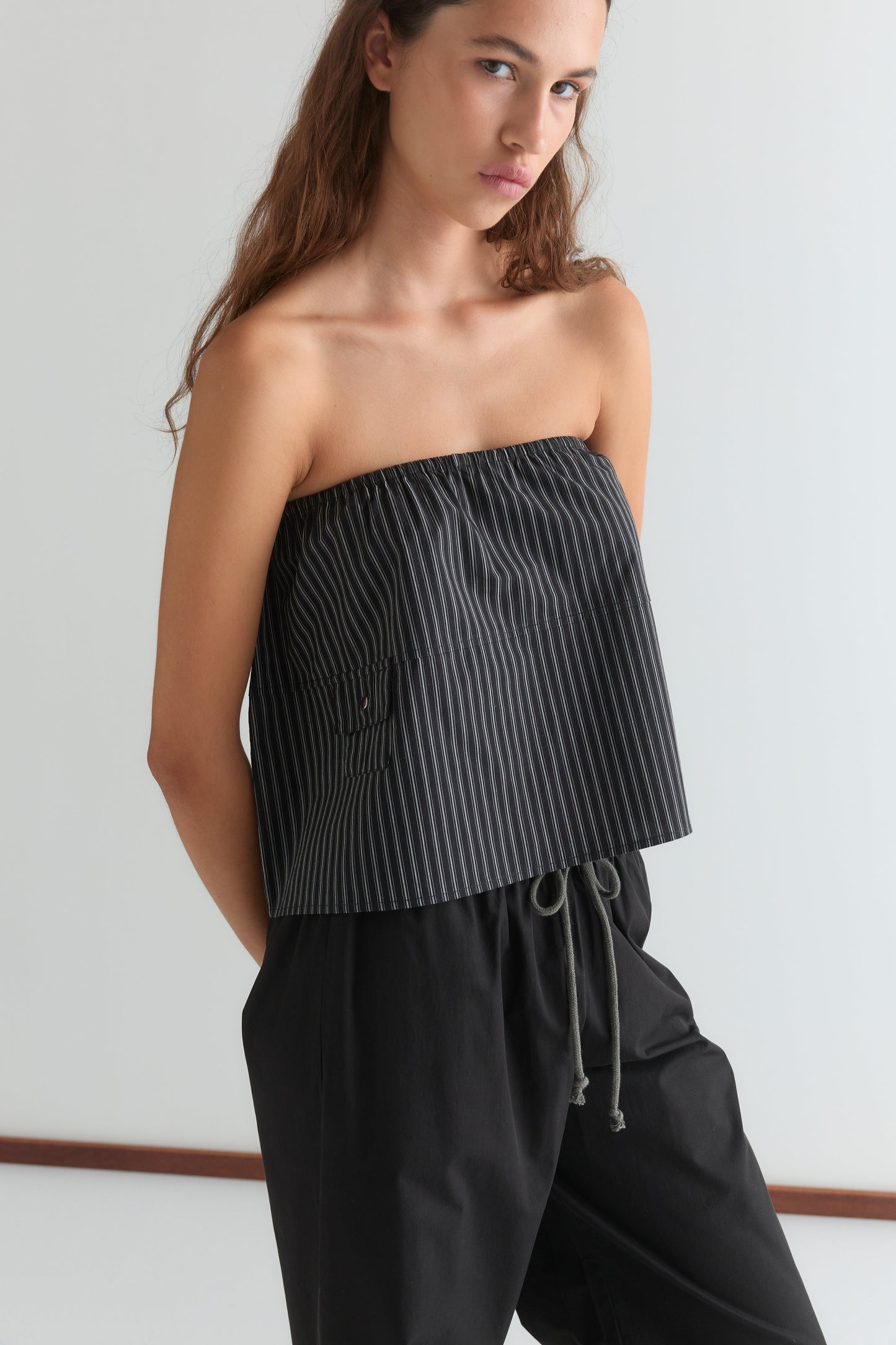 A woman with long brown hair wears the Pocket Skirt - Mono Stripe, a black skirt with thin white stripes and an elasticated waistband, paired with a black strapless top, standing against a plain white background.