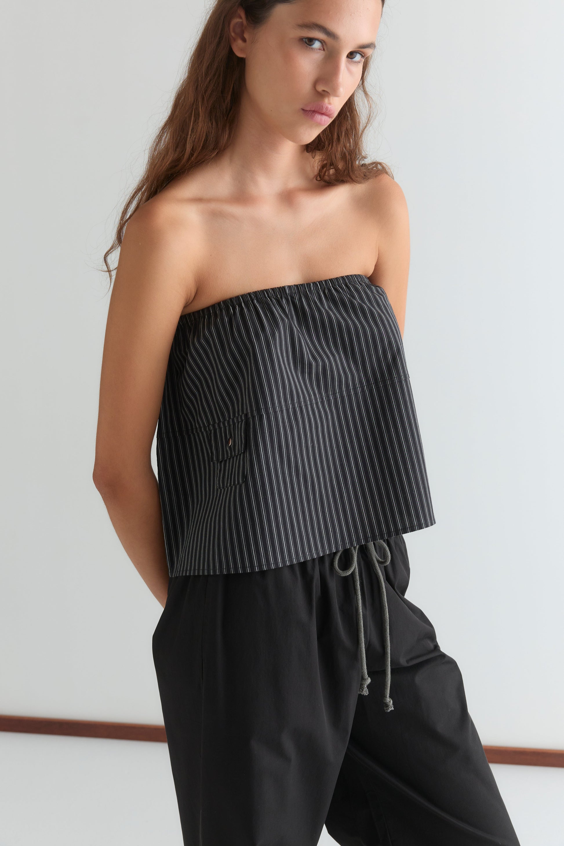 A woman with long brown hair wears the Pocket Skirt - Mono Stripe, a black skirt with thin white stripes and an elasticated waistband, paired with a black strapless top, standing against a plain white background.
