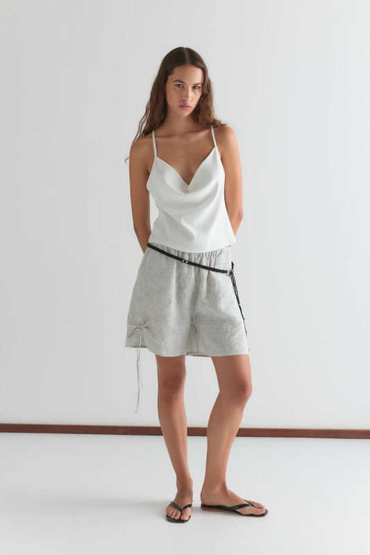 A woman with long brown hair, in a white sleeveless top, Deiji Studios Eyelet Short - Fine Stripe with a black belt, and black flip-flops, stands against a plain white wall.