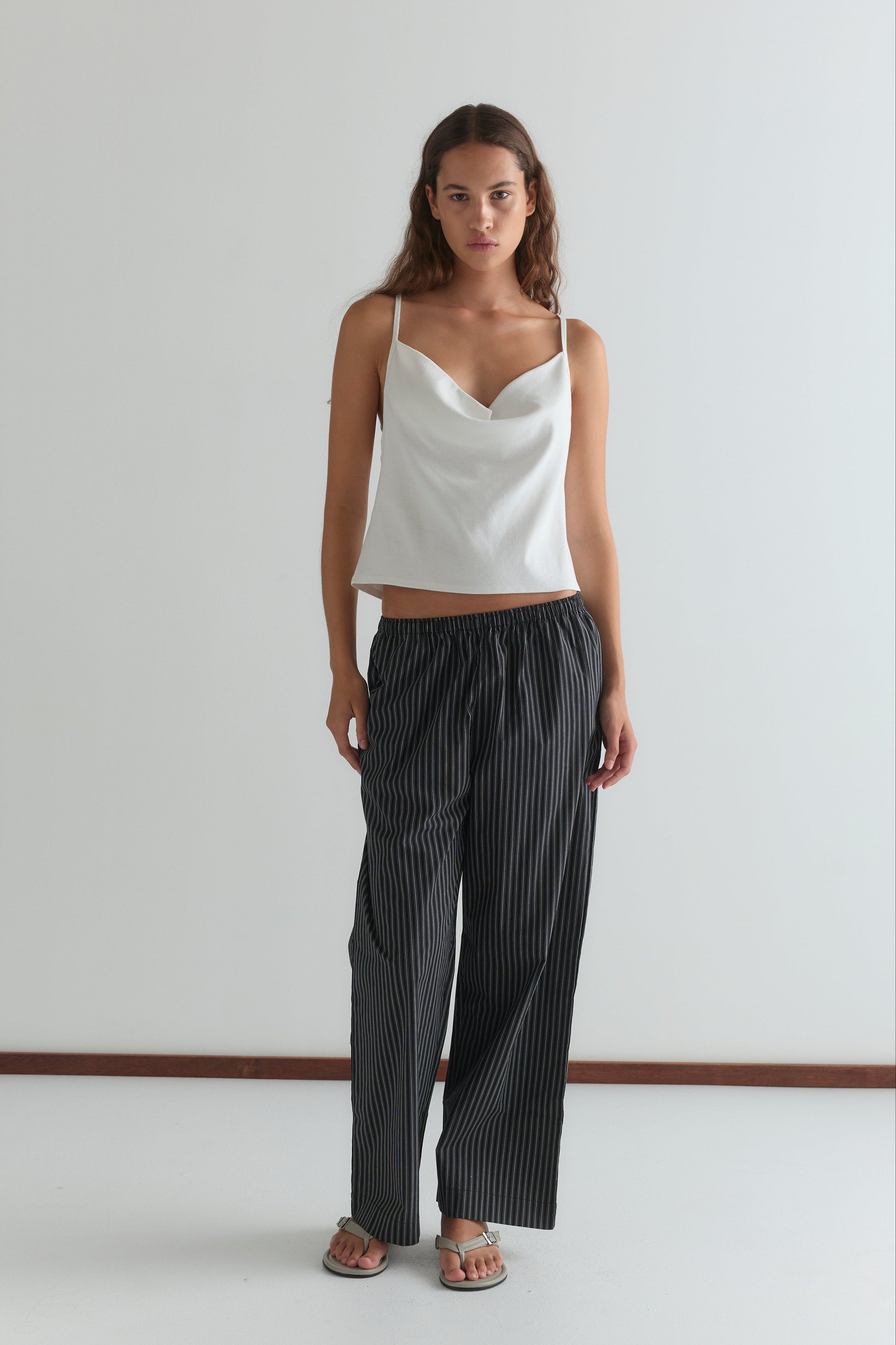 A woman with long brown hair and a neutral expression stands against a white wall wearing a white sleeveless cowl-neck top, sandals, and the Deiji Studios Ease Trouser - Mono Stripe.