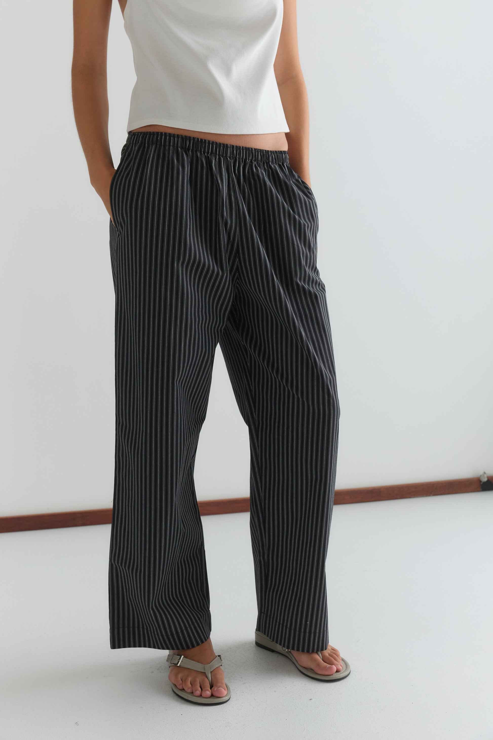A person wearing a white sleeveless top, Ease Trouser - Mono Stripe by Deiji Studios, and sandals stands with hands in their pockets against a plain white background.