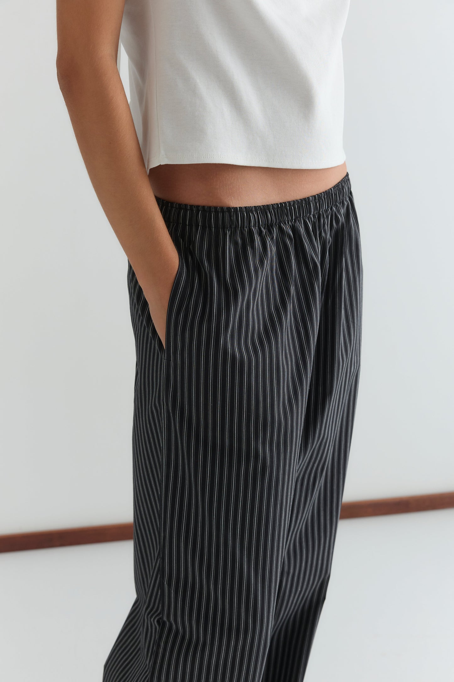 A person wearing a white short-sleeve top and black Deiji Studios Ease Trouser - Mono Stripe stands with one hand in their pocket against a plain background.