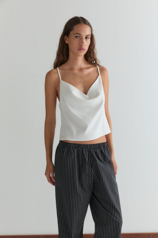 A woman with long brown hair stands against a white wall, wearing a Deiji Studios white sleeveless top and the Ease Trouser - Mono Stripe. She has a neutral expression and her left hand rests at her side.