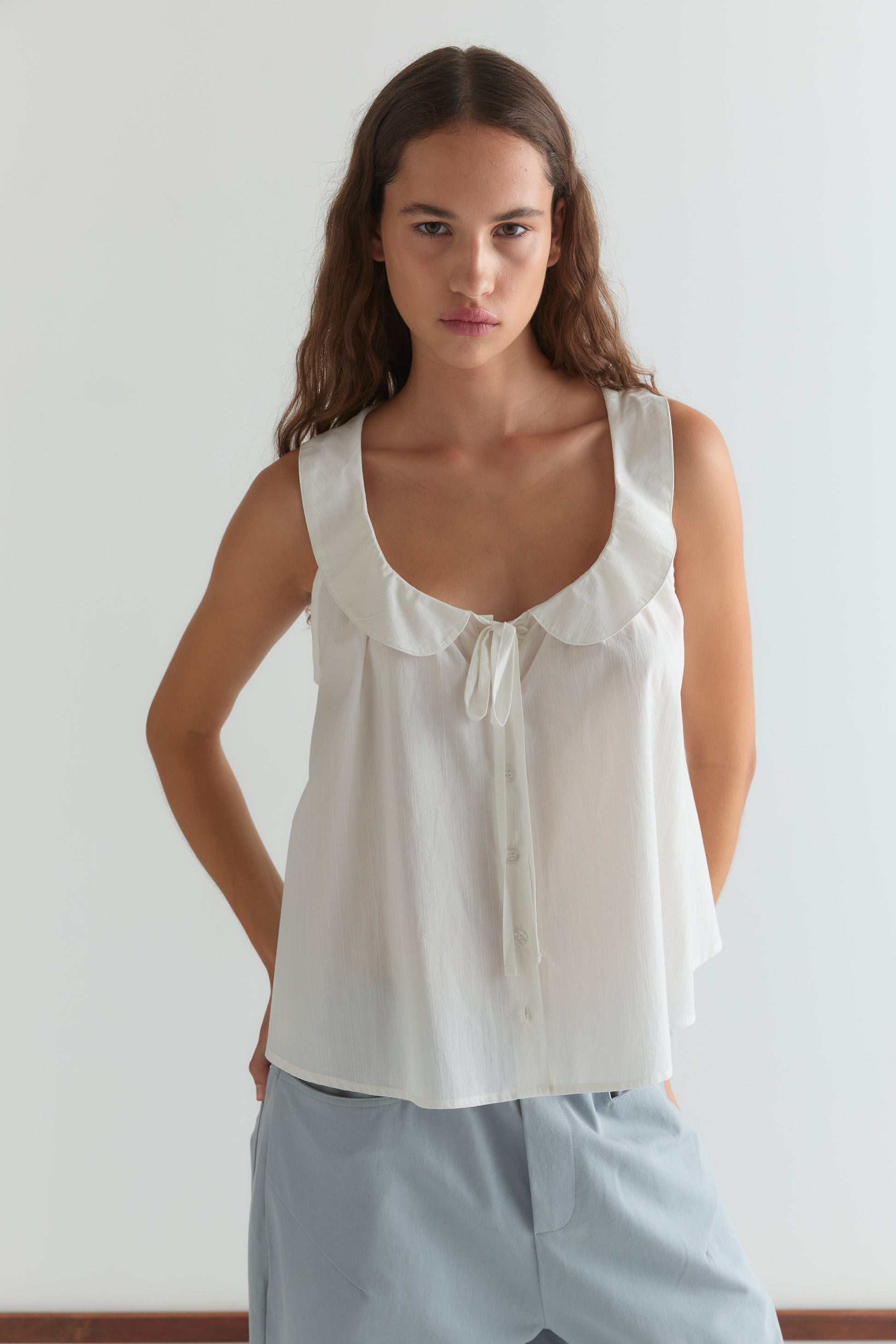 A woman with long brown hair wears the Round Collar Top - White Texture, a sleeveless blouse with a ruffled collar and bow, paired with light blue pants, standing against a plain white background.