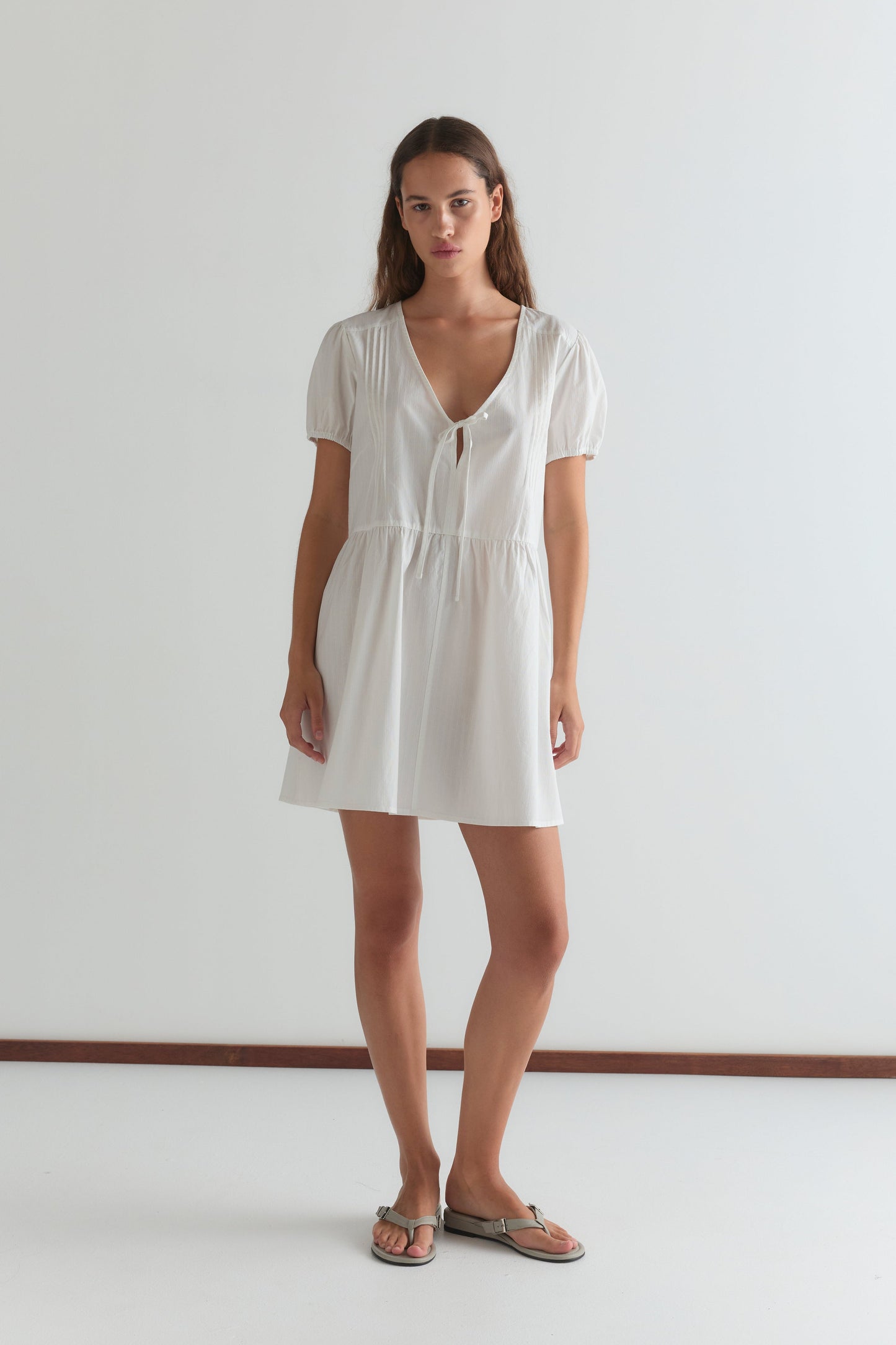 A woman stands indoors against a plain white wall, wearing the V Pleat Dress - White Texture by Deiji Studios. The dress features a V-neck and short puff sleeves. She has long, wavy hair and finishes the look with flat sandals.