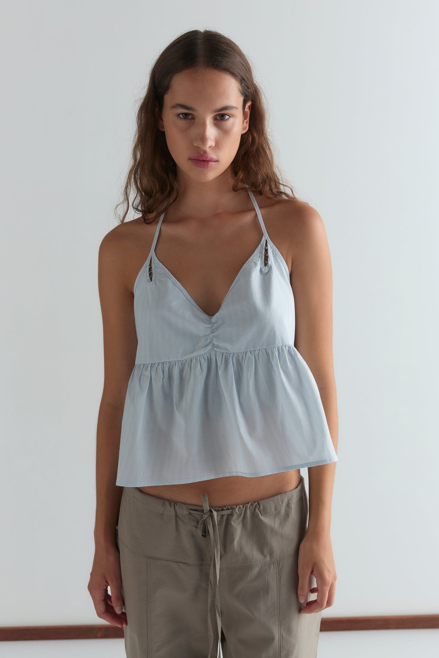 A woman with long brown hair wears the Deiji Studios Keyhole Halter Top in Powder Blue Stripe and khaki drawstring pants, standing against a plain white background.