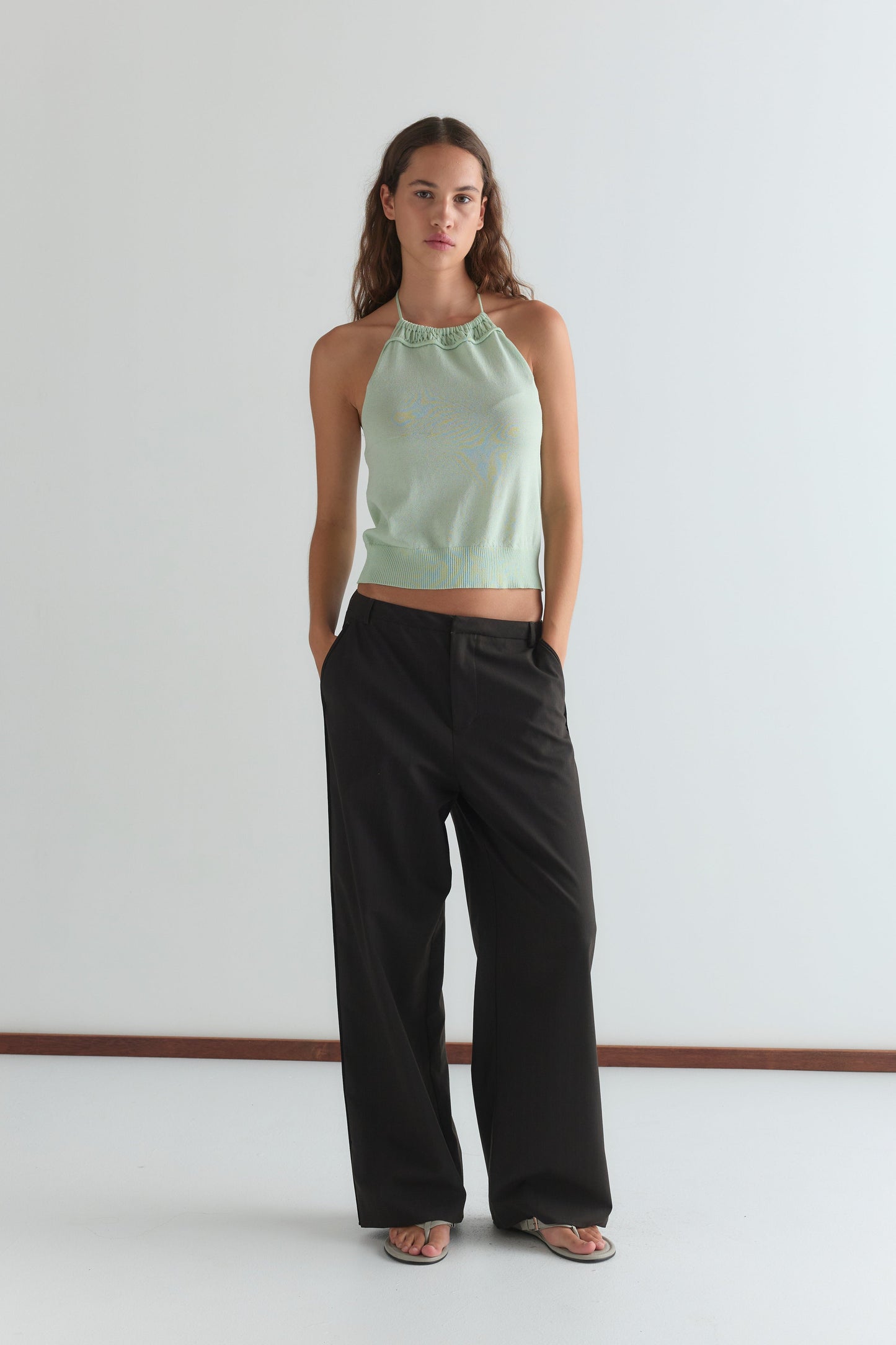 A woman with long, wavy hair stands against a white wall wearing the Deiji Studios Drawcord Knit Top - Mint with an adjustable ruched neckline, loose black pants, and sandals. Her hands are in her pockets.