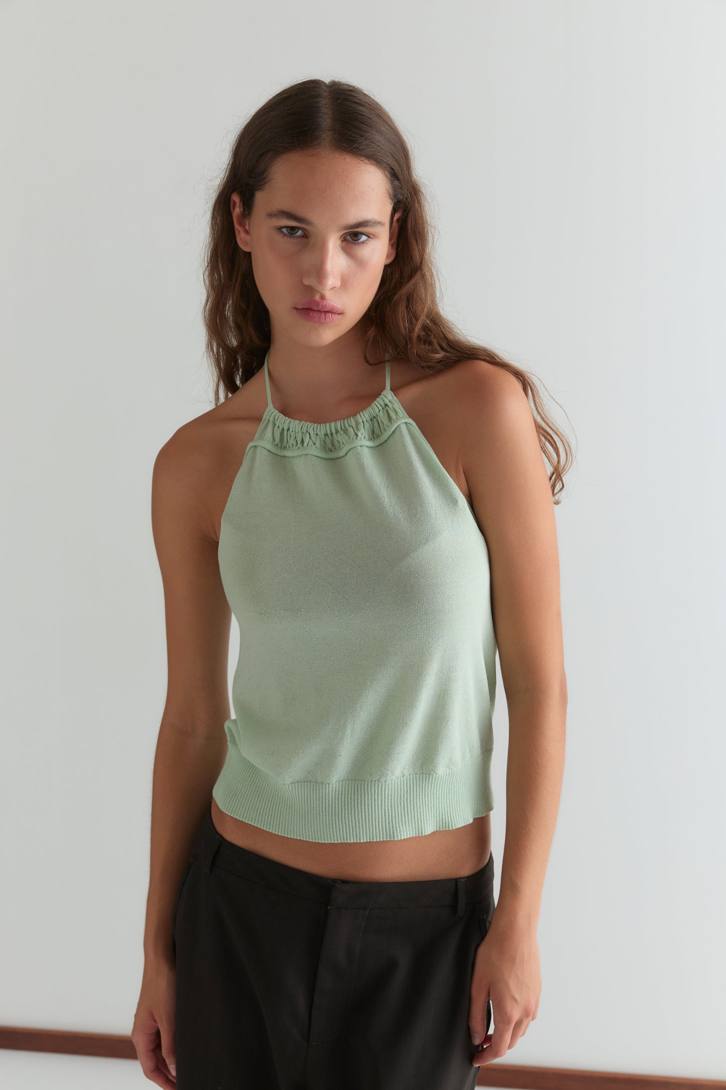A young woman with long brown hair wears the Deiji Studios Drawcord Knit Top in Mint, featuring an adjustable ruched neckline, paired with dark pants. She stands against a white background, looking at the camera with a neutral expression.
