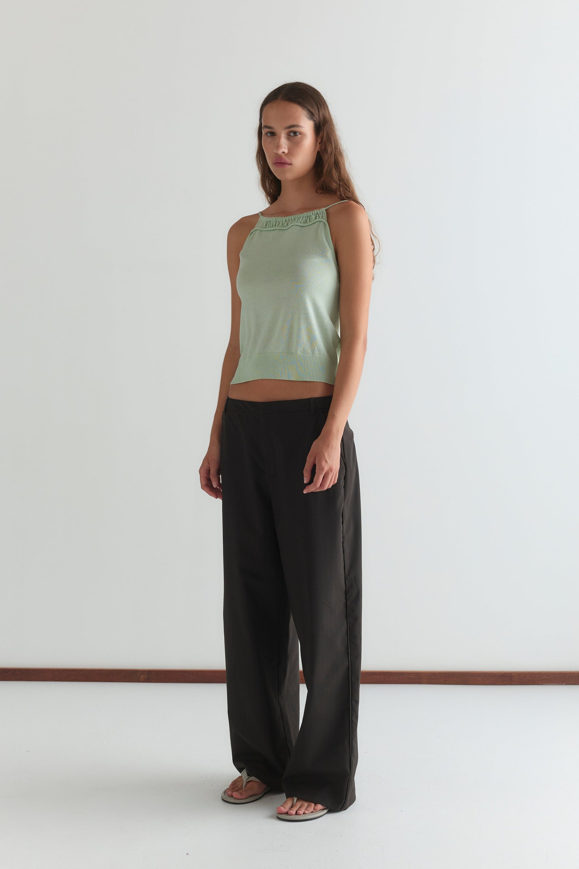 A woman with long brown hair stands indoors against a plain white wall, wearing the Drawcord Knit Top - Mint by Deiji Studios, loose black pants, and white sandals. She has a neutral expression.