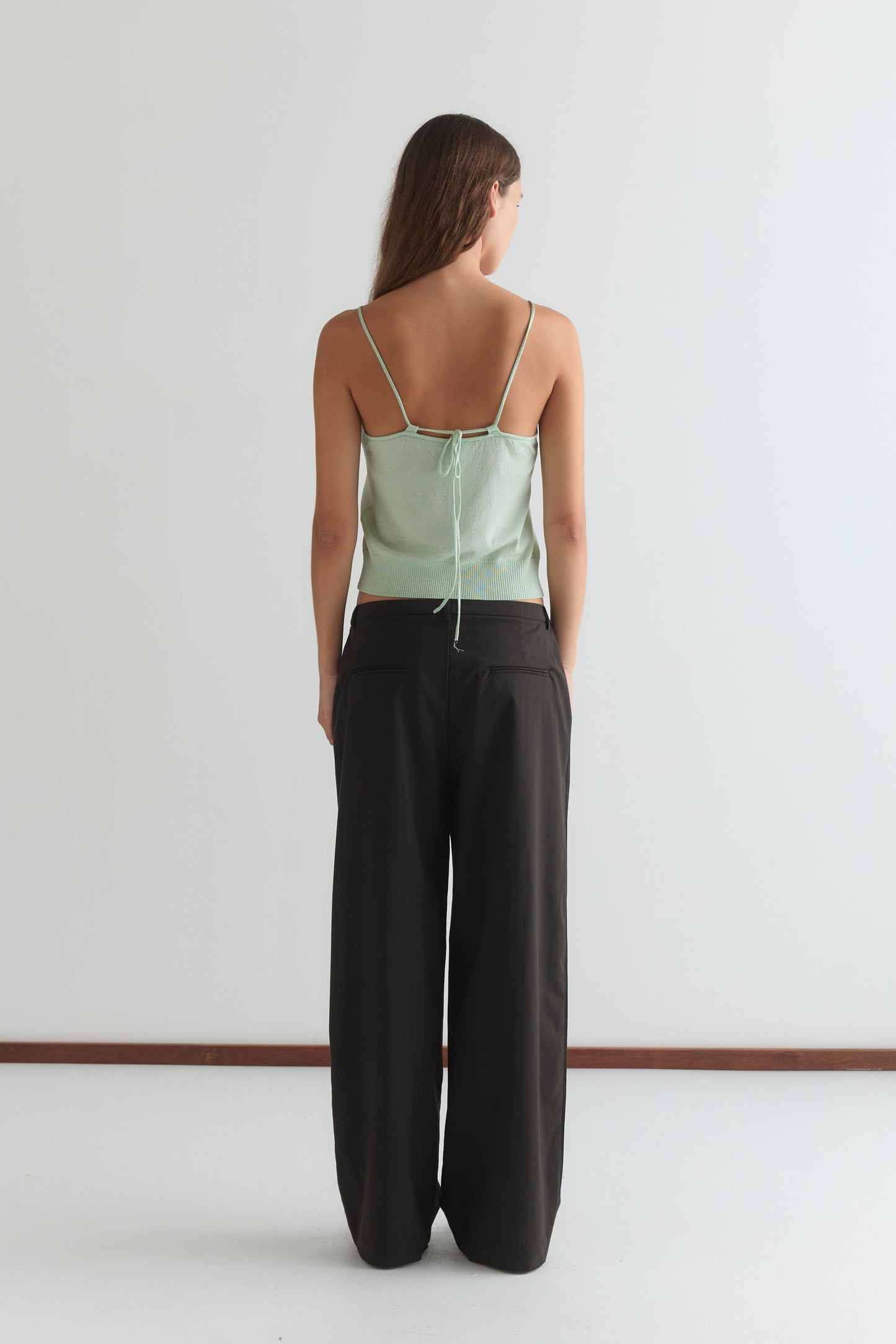 A woman with long brown hair stands facing away from the camera in a mint Deiji Studios Drawcord Knit Top with an adjustable ruched neckline, paired with loose black pants, set against a plain white background.