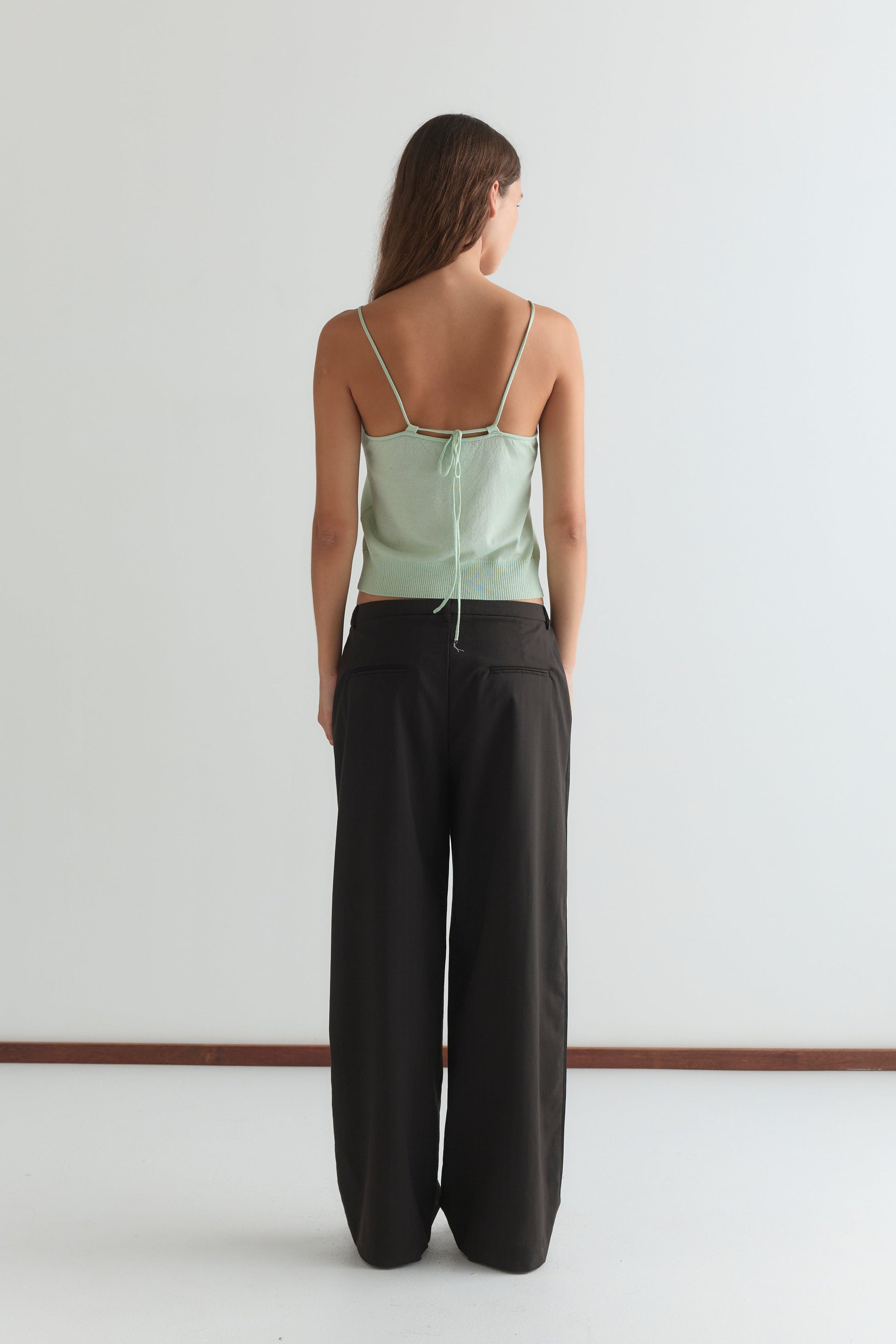 A woman with long brown hair stands facing away from the camera in a mint Deiji Studios Drawcord Knit Top with an adjustable ruched neckline, paired with loose black pants, set against a plain white background.