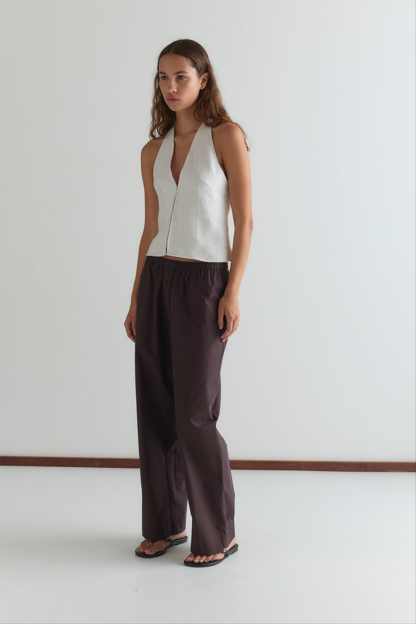 A woman stands against a plain white wall, wearing the Close Halter Top - White from Deiji Studios, loose dark brown pants, and black sandals. She has long, wavy hair and a neutral expression.