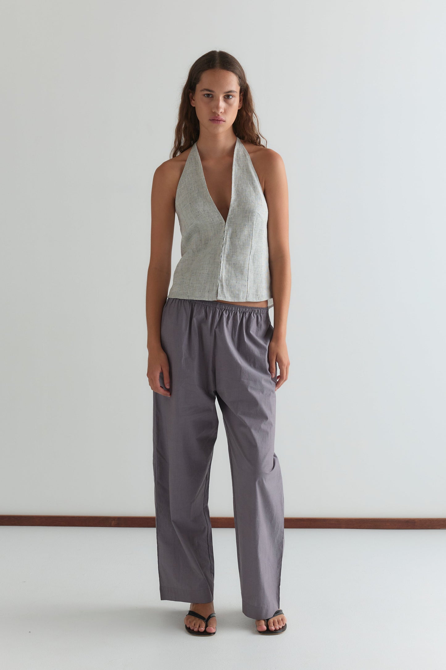 A woman with long brown hair and black sandals stands against a plain white wall, wearing Deiji Studios' Close Halter Top - Fine Stripe with a deep V neckline and loose, high-waisted gray pants.