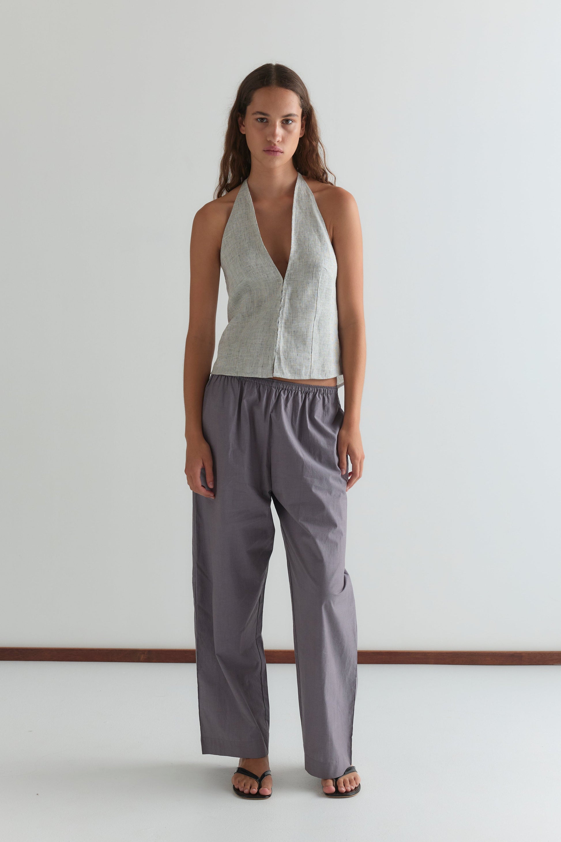 A woman with long brown hair and black sandals stands against a plain white wall, wearing Deiji Studios' Close Halter Top - Fine Stripe with a deep V neckline and loose, high-waisted gray pants.