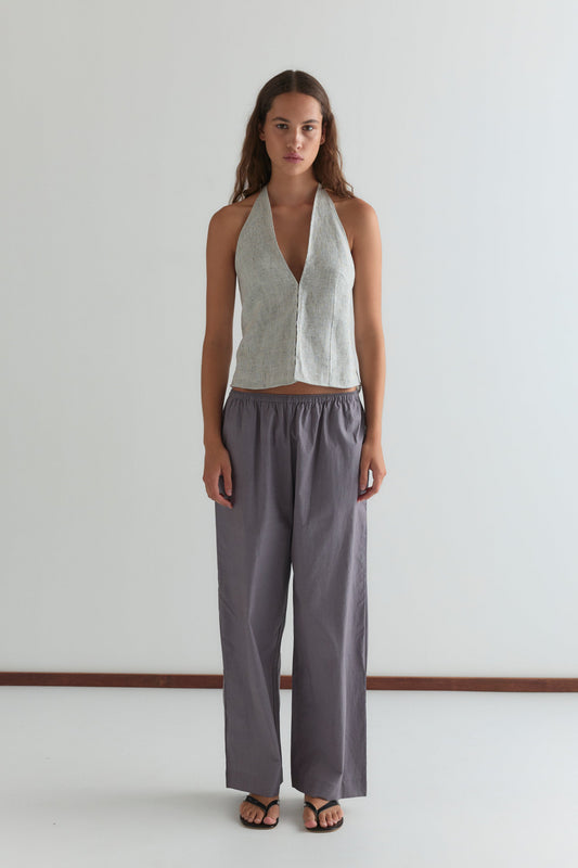 A woman with long brown hair and a neutral expression stands against a white wall, wearing the Ease Trouser—loose, high-waisted gray pants—paired with a sleeveless light gray halter top and black sandals.