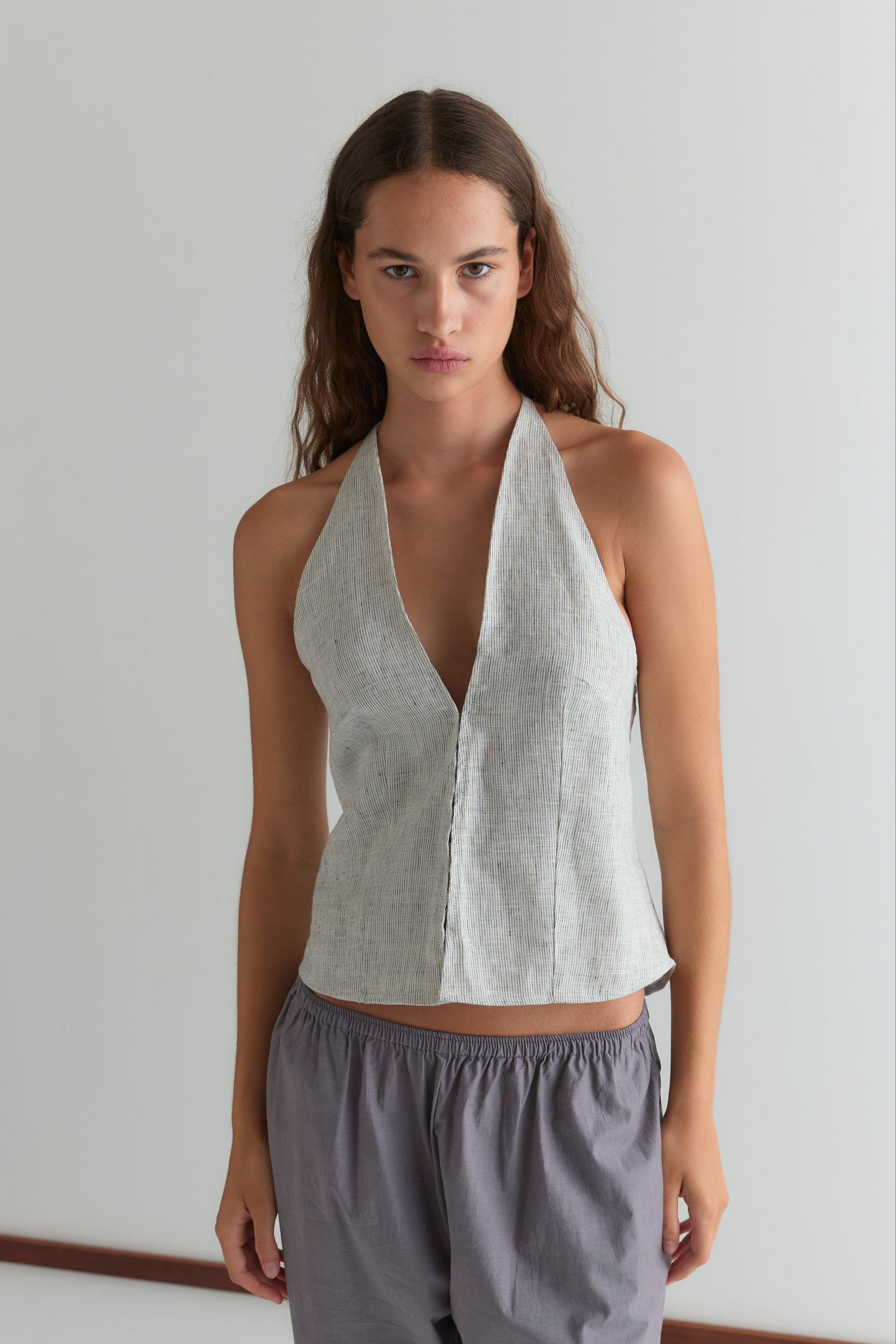 A person with long brown hair stands against a plain white wall, wearing the Close Halter Top - Fine Stripe by Deiji Studios and loose gray pants, looking directly at the camera with a neutral expression.