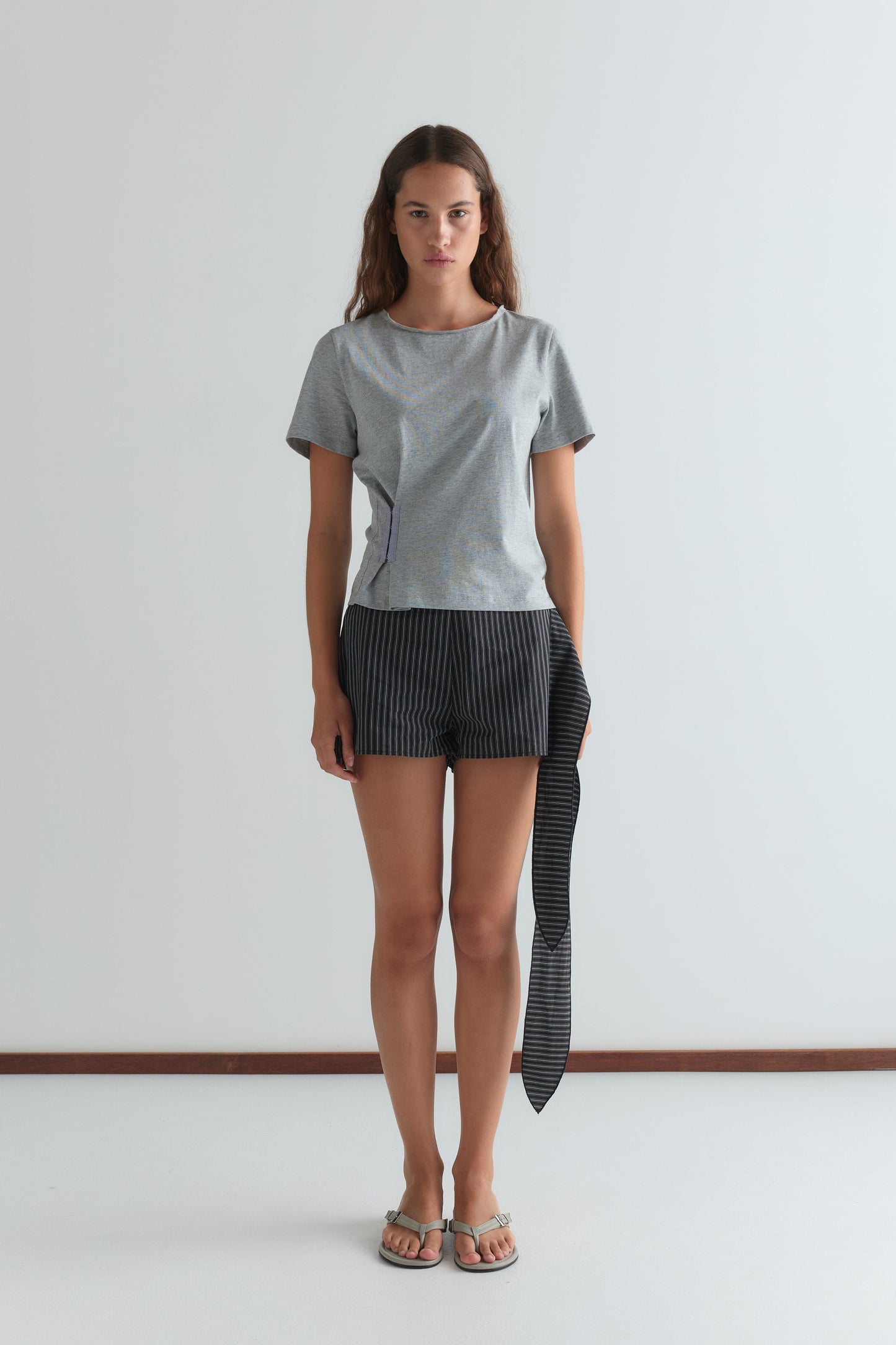 A woman stands against a white wall, wearing a Deiji Studios light gray short-sleeve top, Mono Stripe Belted Short with an attached long fabric detail, and sandals. She faces forward with a neutral expression.