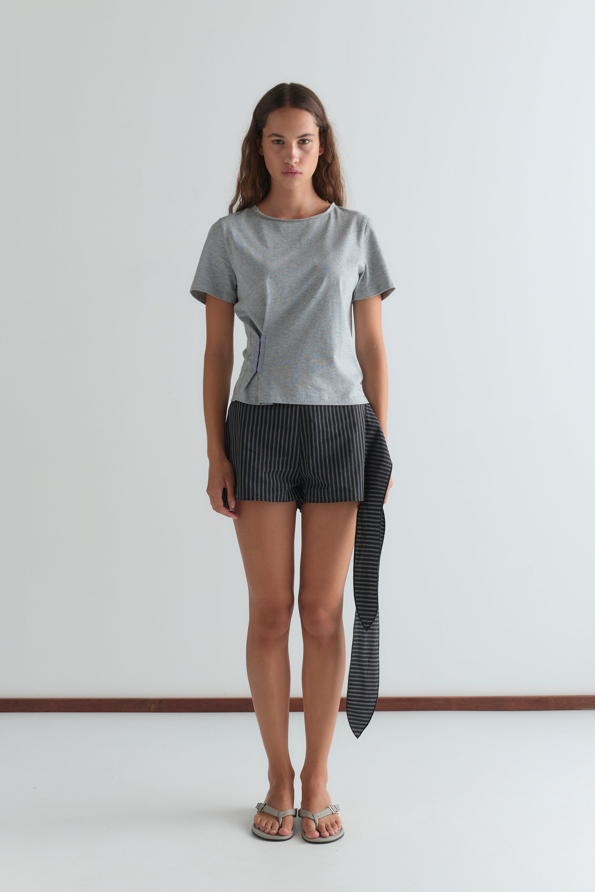 A woman stands against a white wall, wearing a Deiji Studios light gray short-sleeve top, Mono Stripe Belted Short with an attached long fabric detail, and sandals. She faces forward with a neutral expression.