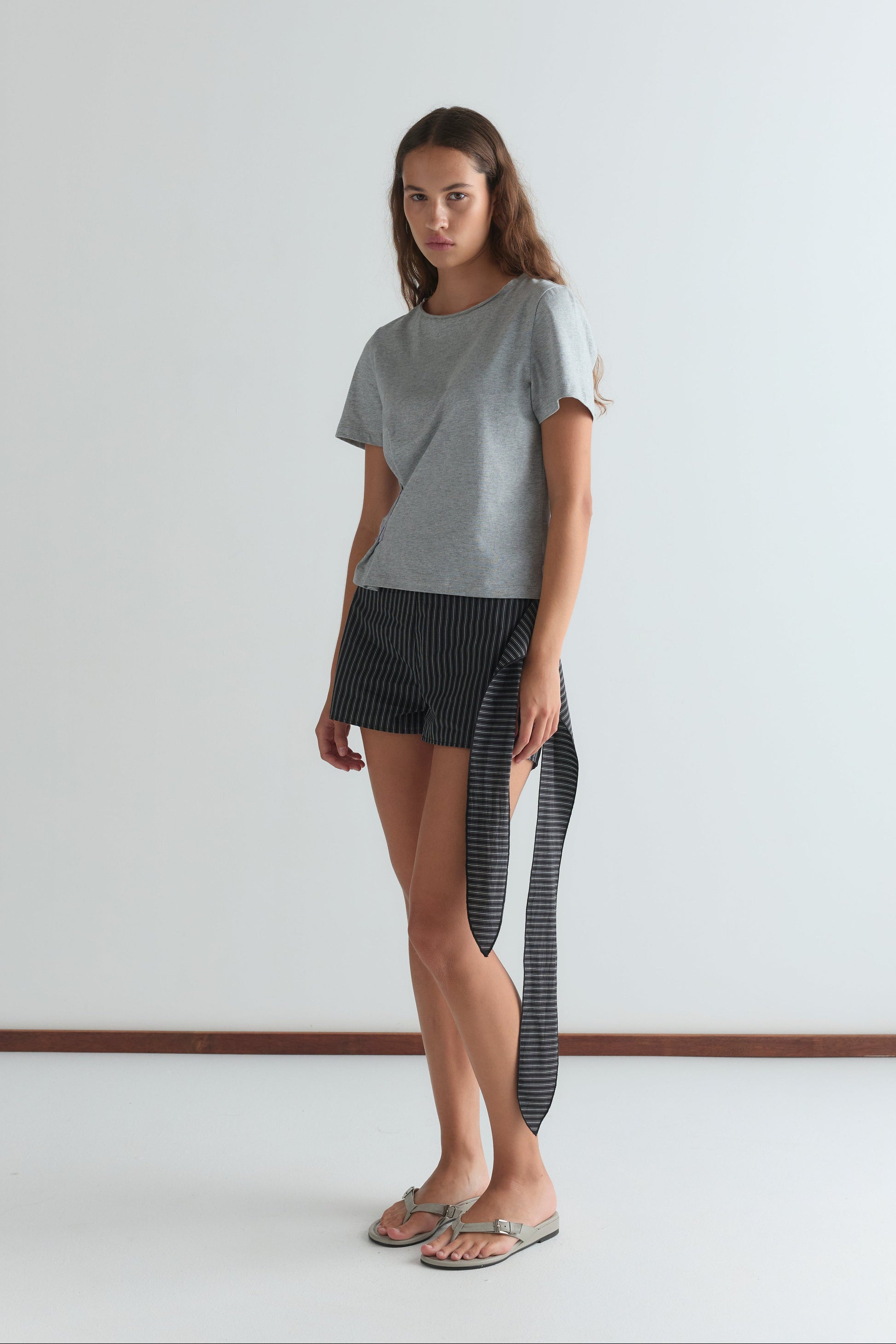 A woman with long brown hair stands indoors against a plain white wall, wearing a gray t-shirt, Deiji Studios Belted Short - Mono Stripe with a long side tie, and beige flip-flops.