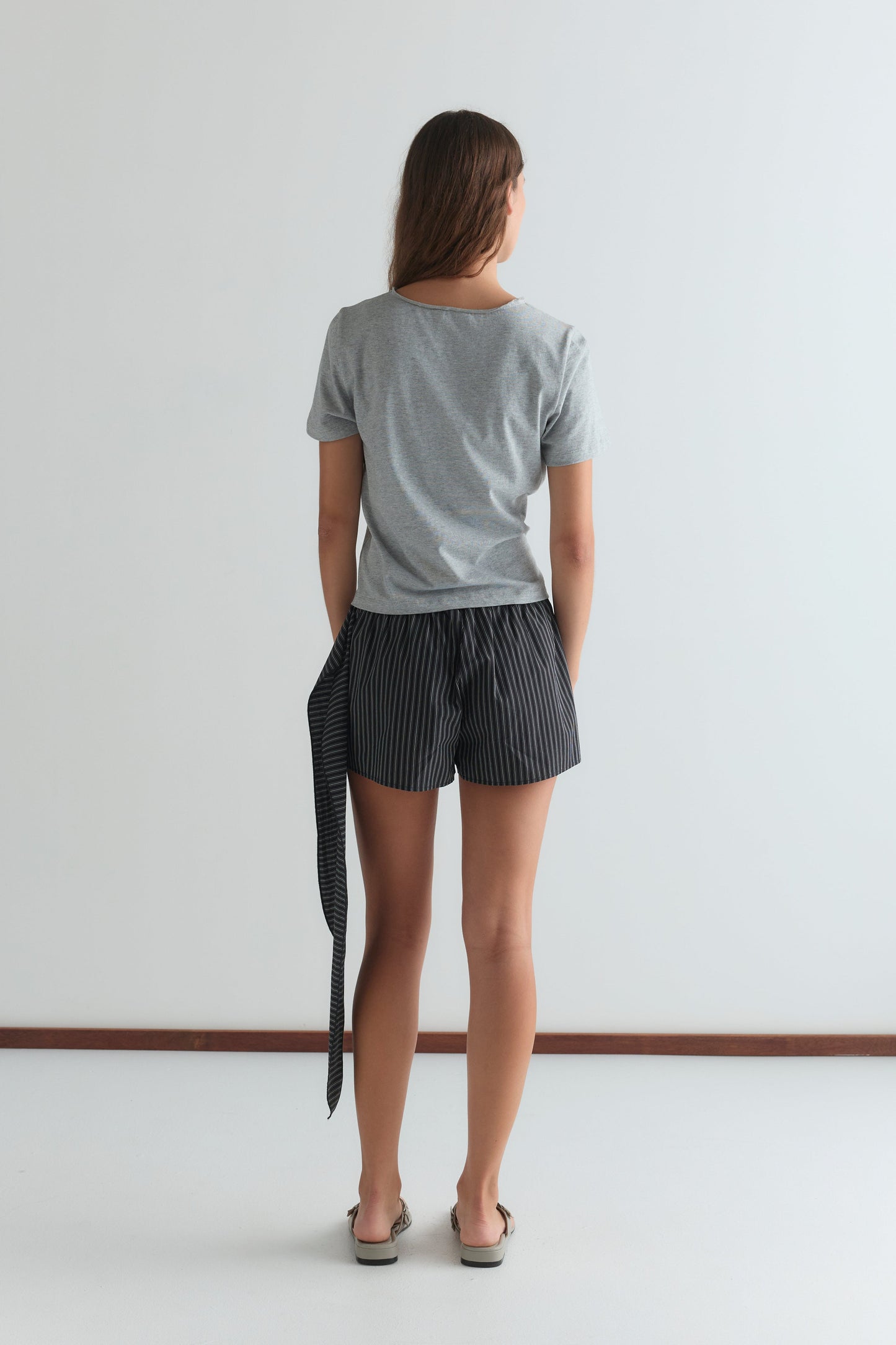 A woman stands facing away, wearing a gray T-shirt and the Belted Short - Mono Stripe by Deiji Studios, featuring black and white stripes and a fabric belt, with her hands in her pockets against a plain white background.