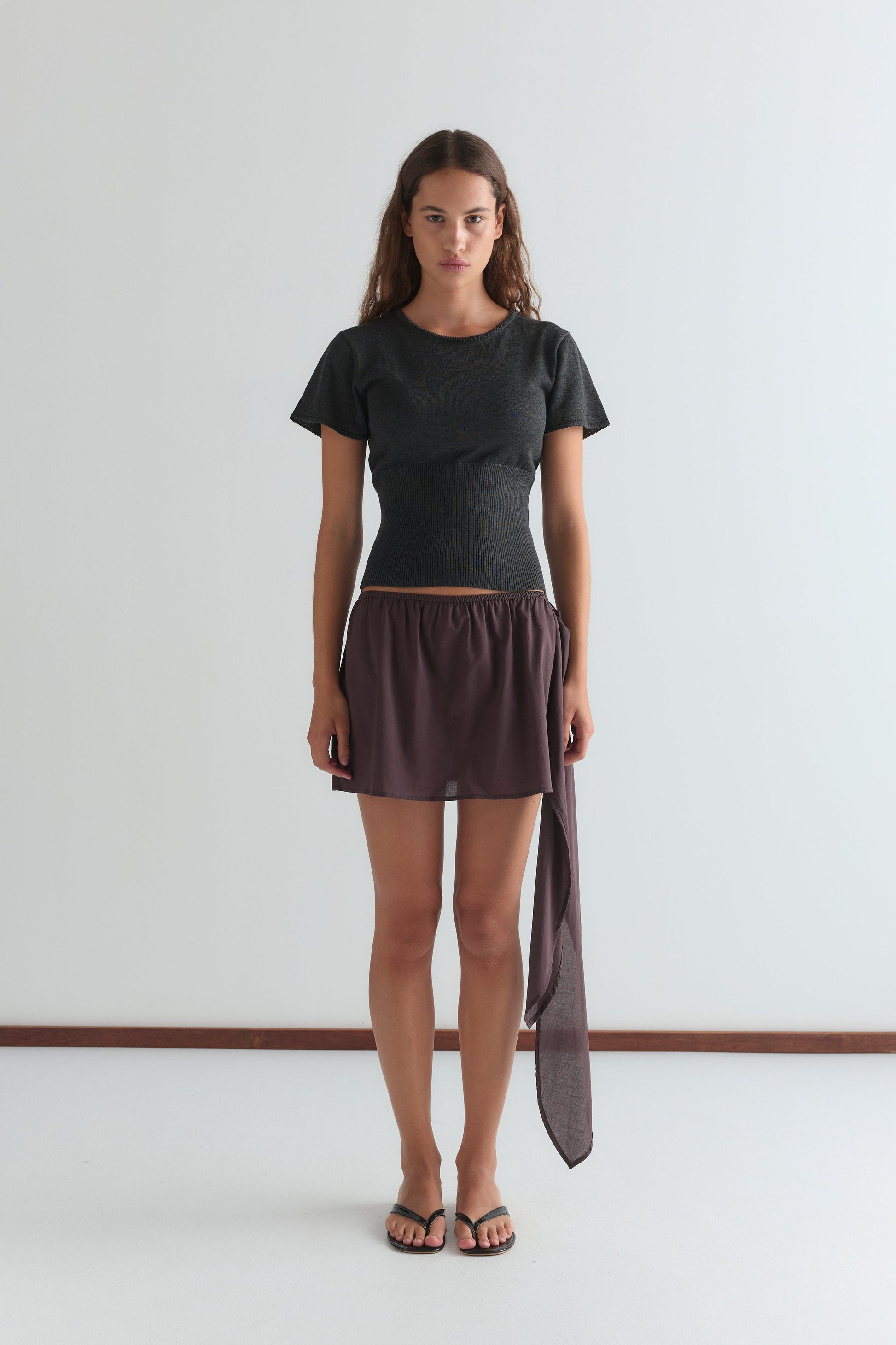 A woman stands against a plain white wall, wearing Deiji Studios' Trim Knit Tee in dark grey, a short brown skirt with an asymmetrical draped detail, and black sandals. She looks forward with a neutral expression.
