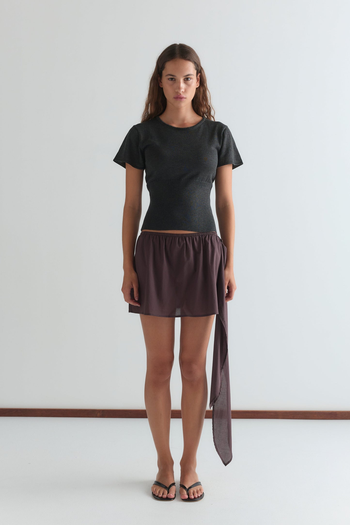 A woman with long brown hair and a neutral expression stands against a plain white wall, wearing a fitted dark gray short-sleeved top, the Belted Skirt - Wine featuring an asymmetrical drape, and black flip-flops.