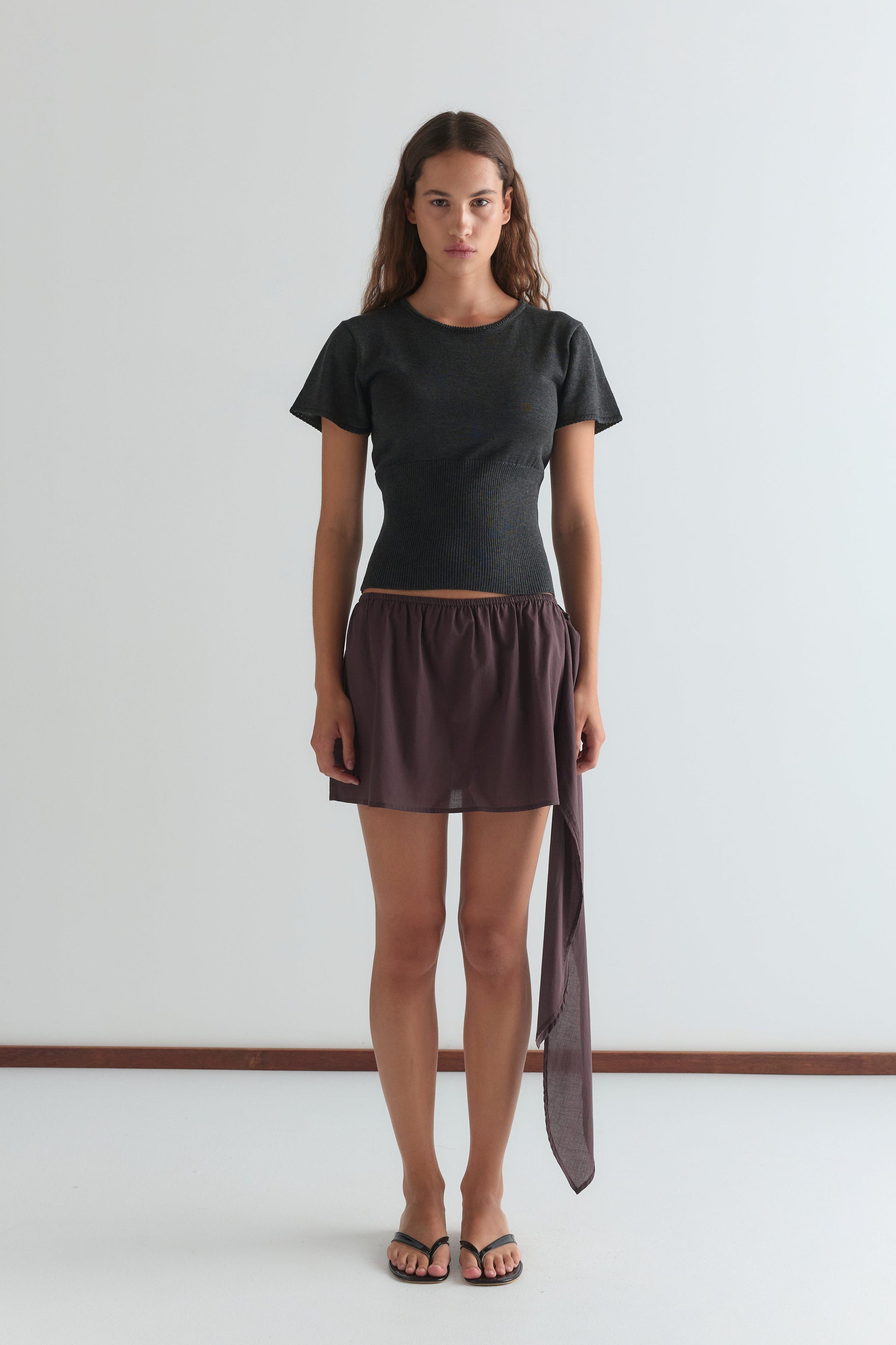 A woman with long brown hair and a neutral expression stands against a plain white wall, wearing a fitted dark gray short-sleeved top, the Belted Skirt - Wine featuring an asymmetrical drape, and black flip-flops.