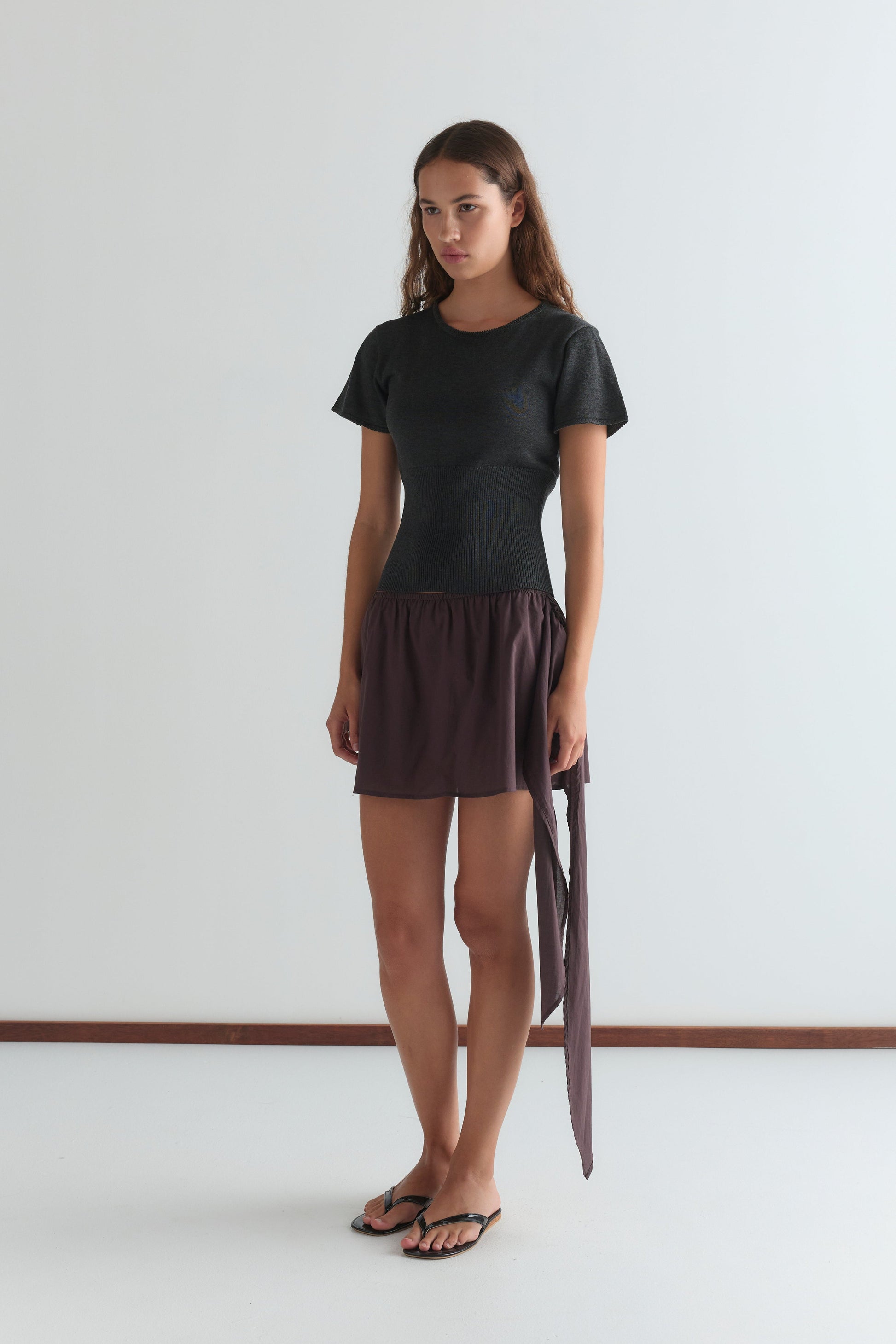A woman with long brown hair wears the Trim Knit Tee in dark grey, a short purple skirt with a long tie detail, and black flip-flops, standing against a plain white background.