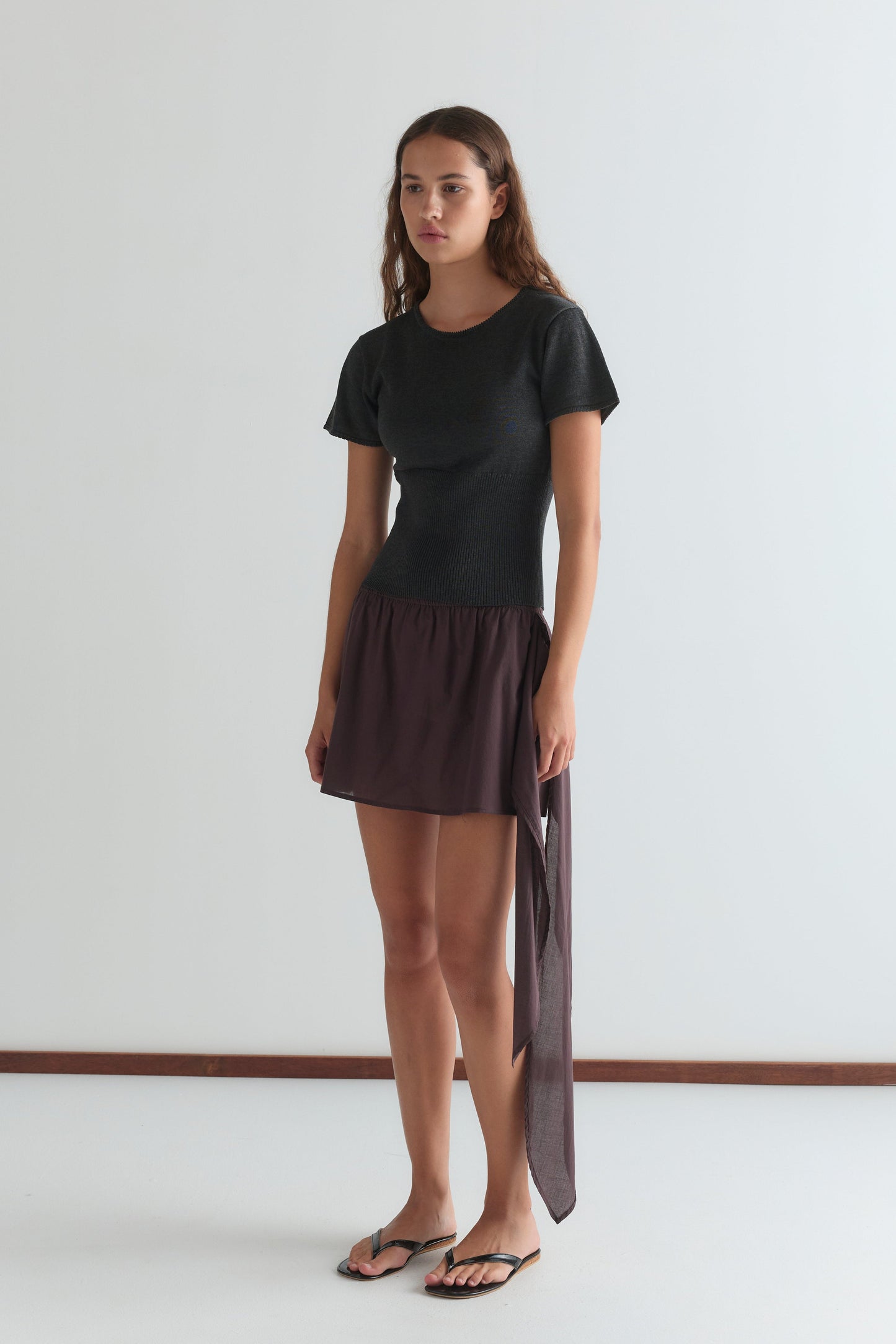 A young woman with long brown hair stands indoors against a white wall, wearing a fitted black short-sleeve top, black sandals, and the Belted Skirt - Wine mini skirt from Deiji Studios.