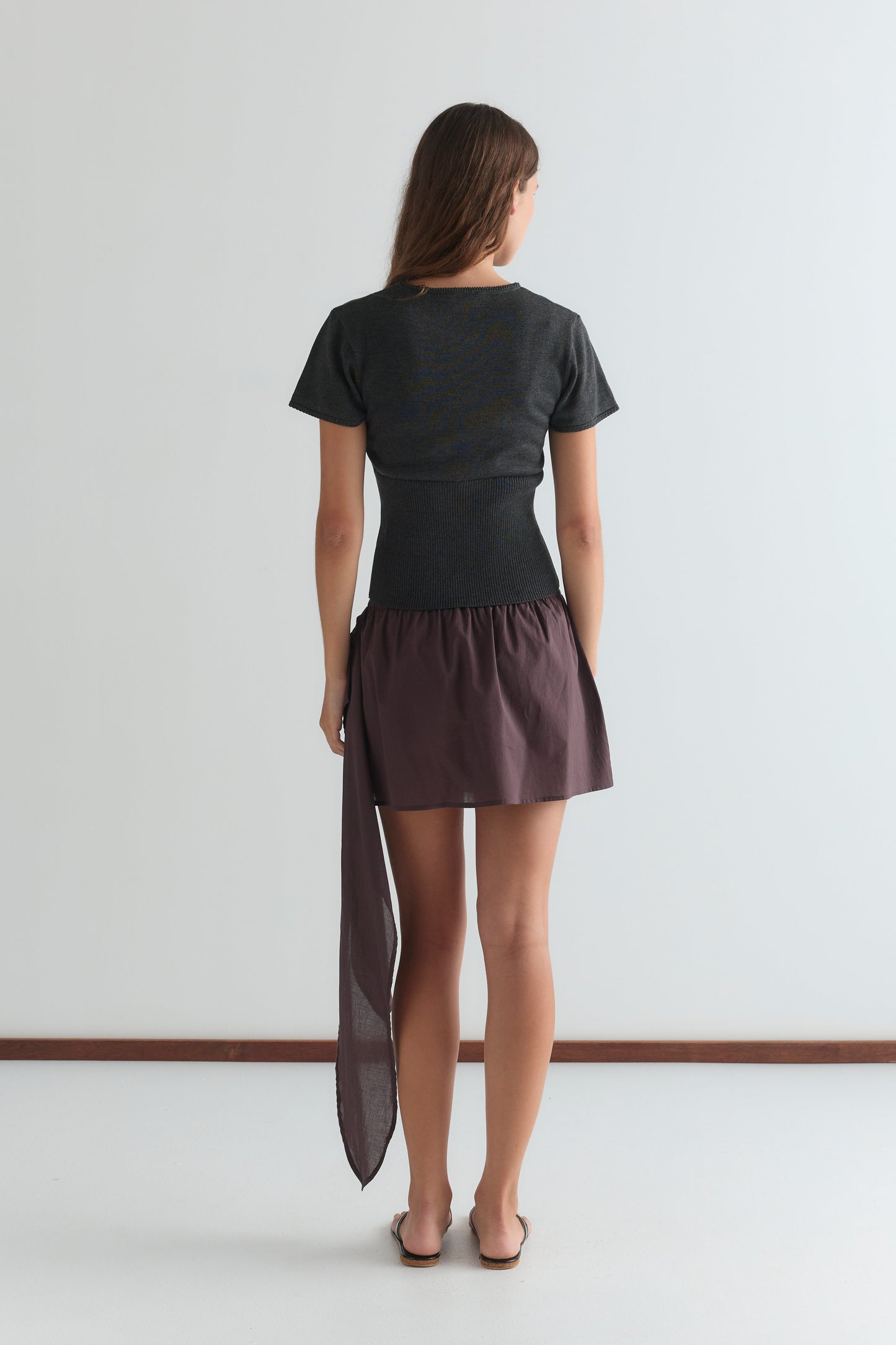 A woman faces away from the camera in a short-sleeved dark top and the Belted Skirt - Wine by Deiji Studios, featuring an asymmetrical sheer detail. She stands barefoot on a white floor against a plain light wall.