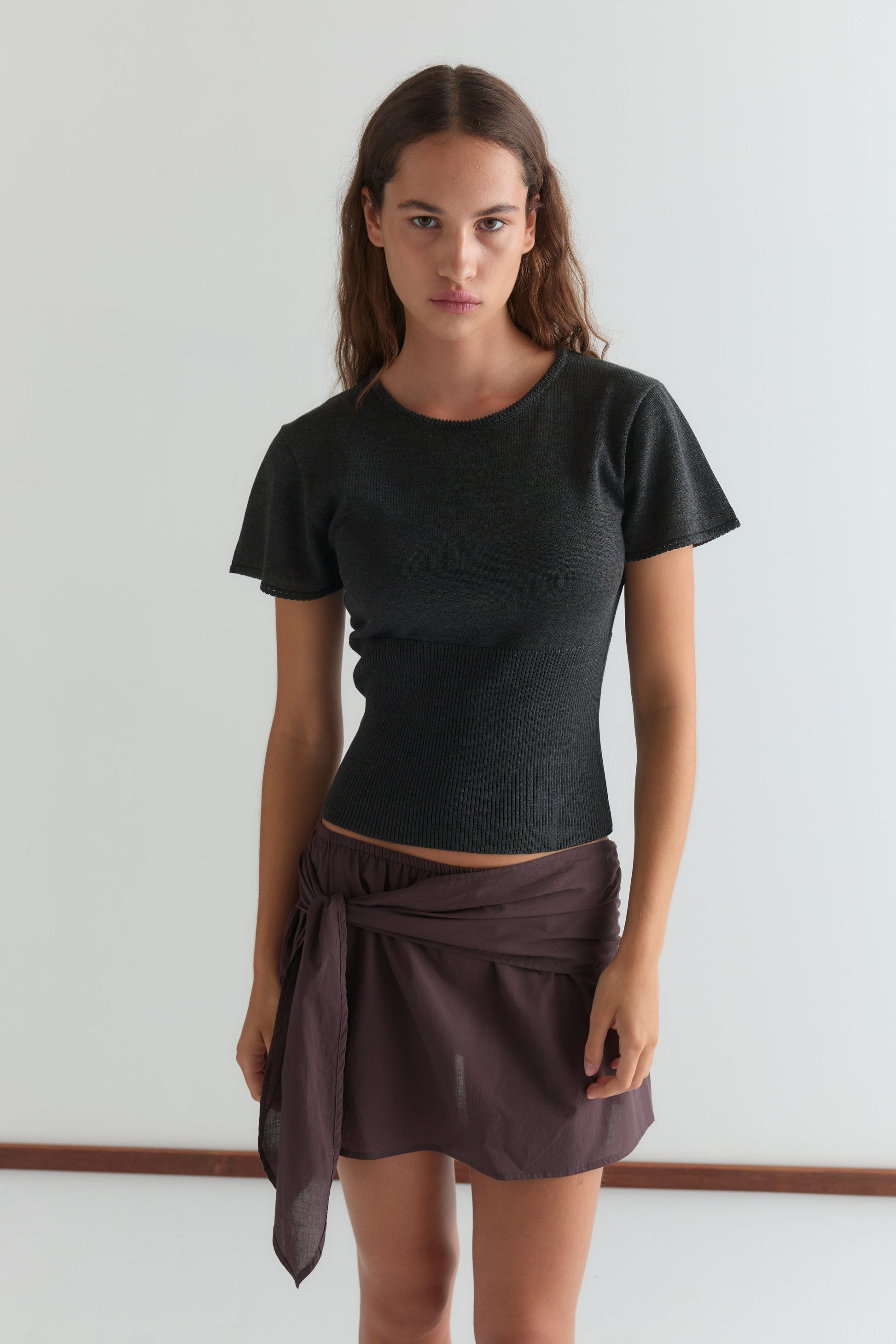 A woman with long brown hair wears a fitted black short-sleeve top and a wine-colored belted mini skirt (Belted Skirt - Wine), standing against a plain light background and looking directly at the camera with a neutral expression.