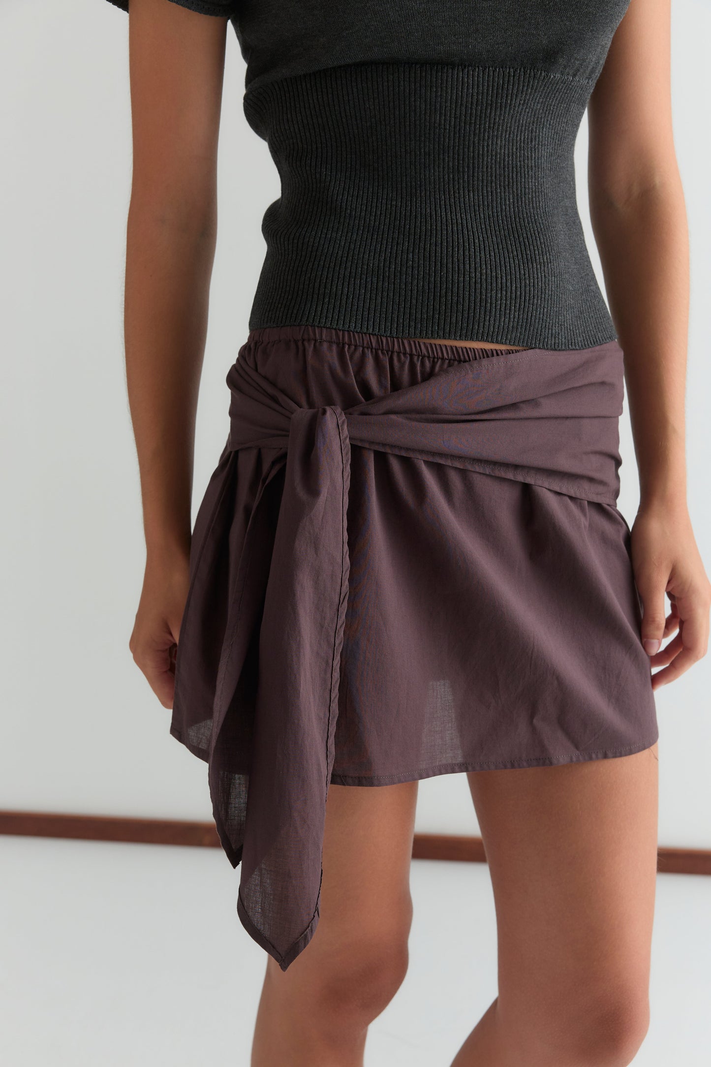 A woman stands indoors against a plain light background, wearing a black ribbed top paired with the Belted Skirt - Wine, which features a short length and side tie detail.