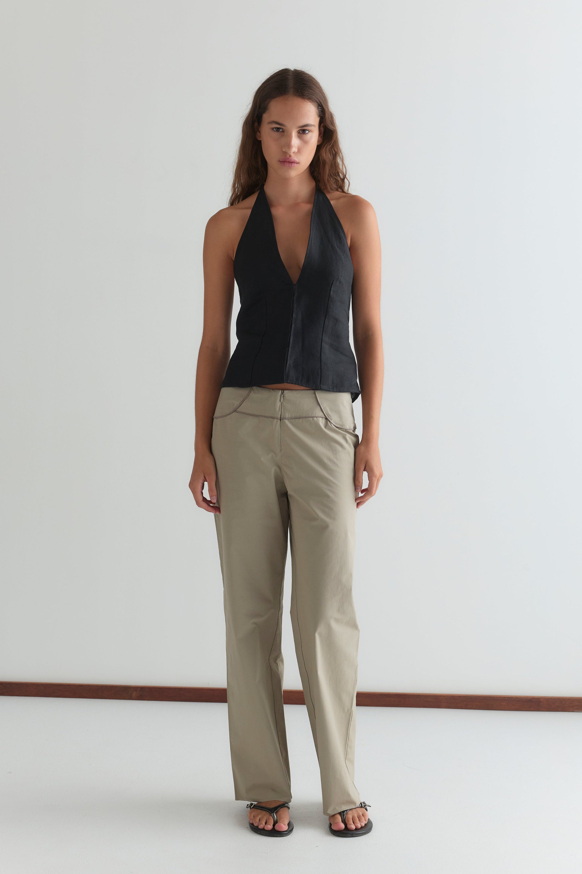 A woman with long brown hair and a neutral expression stands against a white wall, wearing the Close Halter Top - Black from Deiji Studios, beige zip-detail trousers, and black sandals.