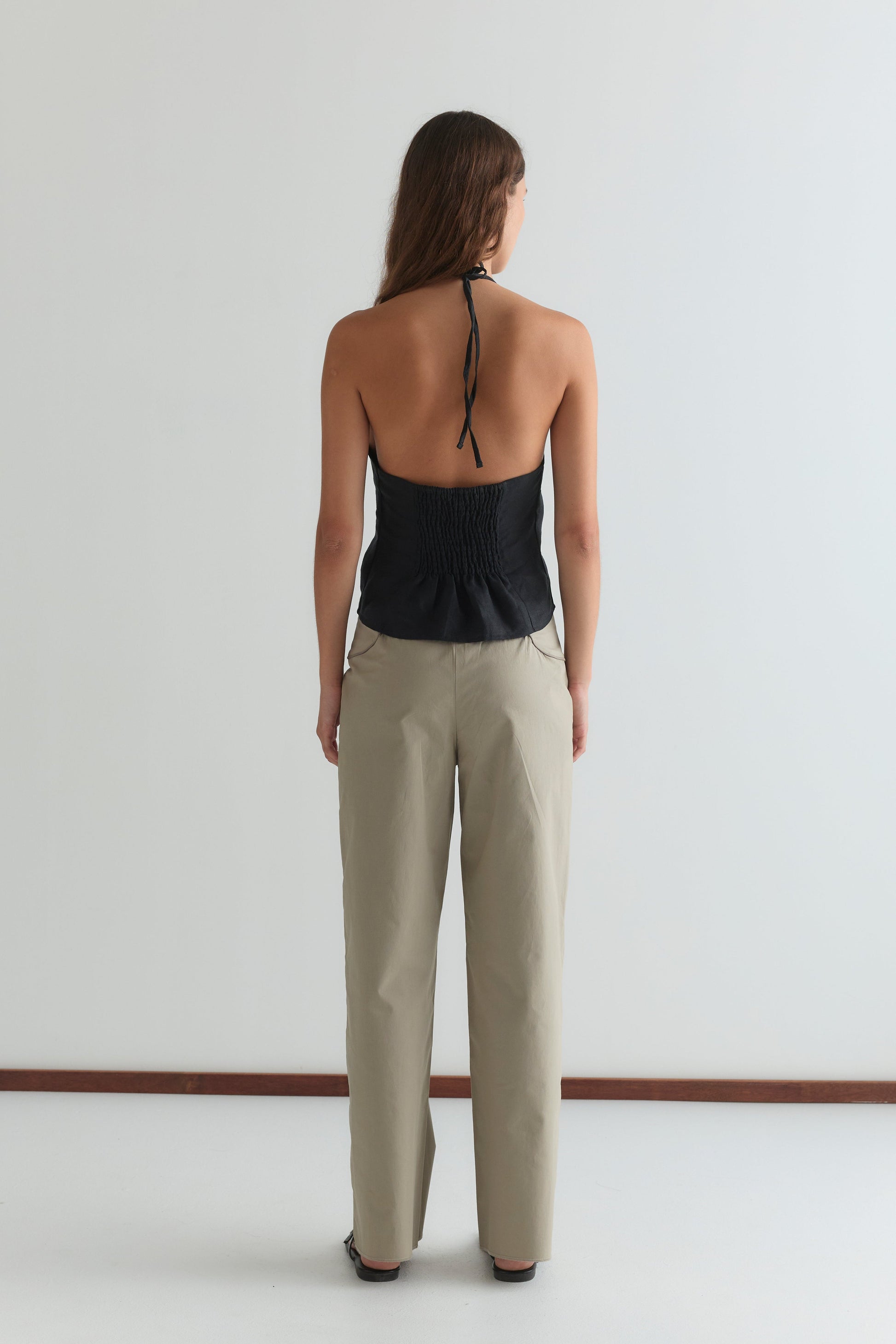 A woman faces away from the camera, wearing the Close Halter Top - Black by Deiji Studios with an open back and beige pants against a plain white background.