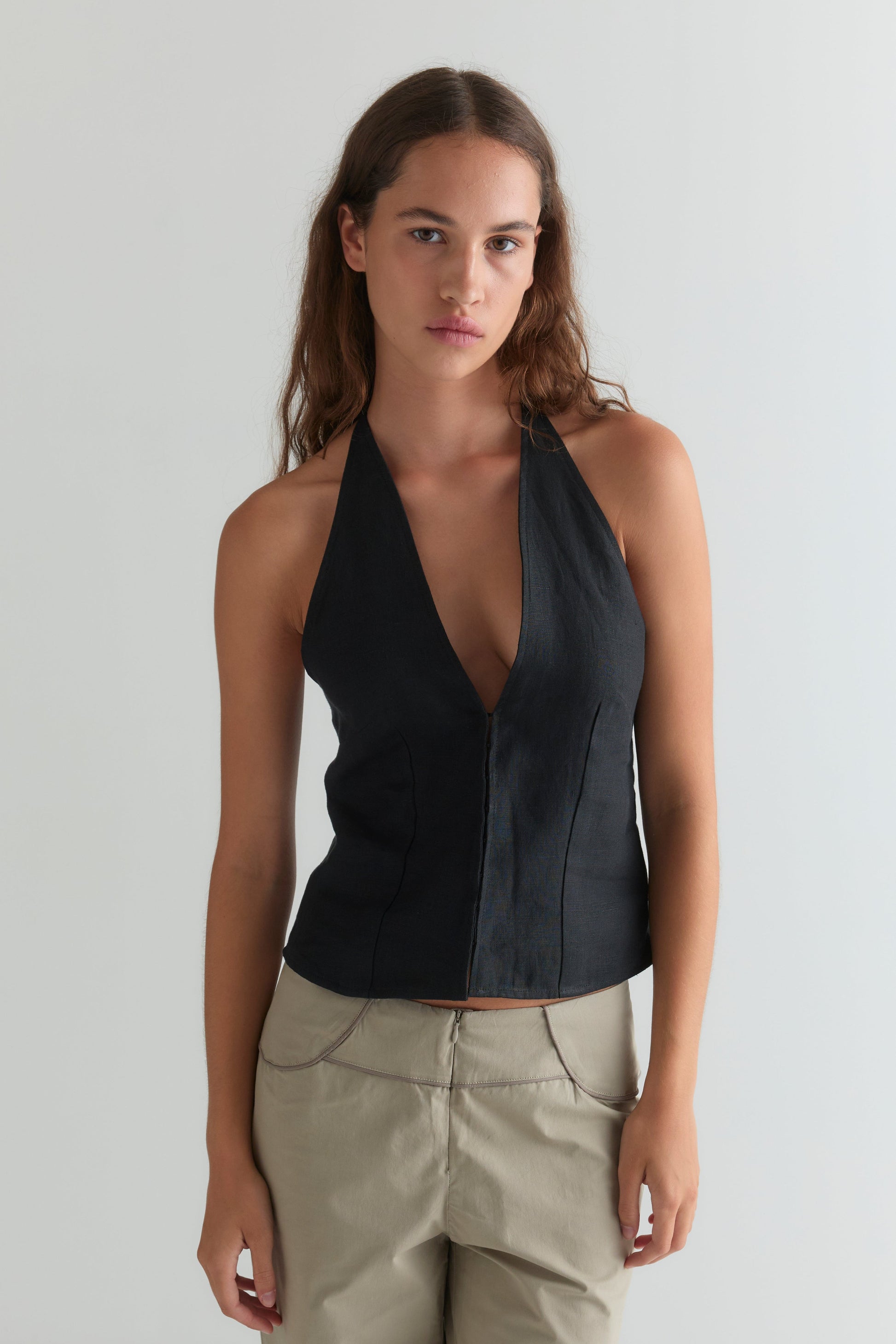 A woman with long brown hair stands against a plain light background, wearing the Close Halter Top - Black by Deiji Studios and light beige pants. She faces forward with a neutral expression.
