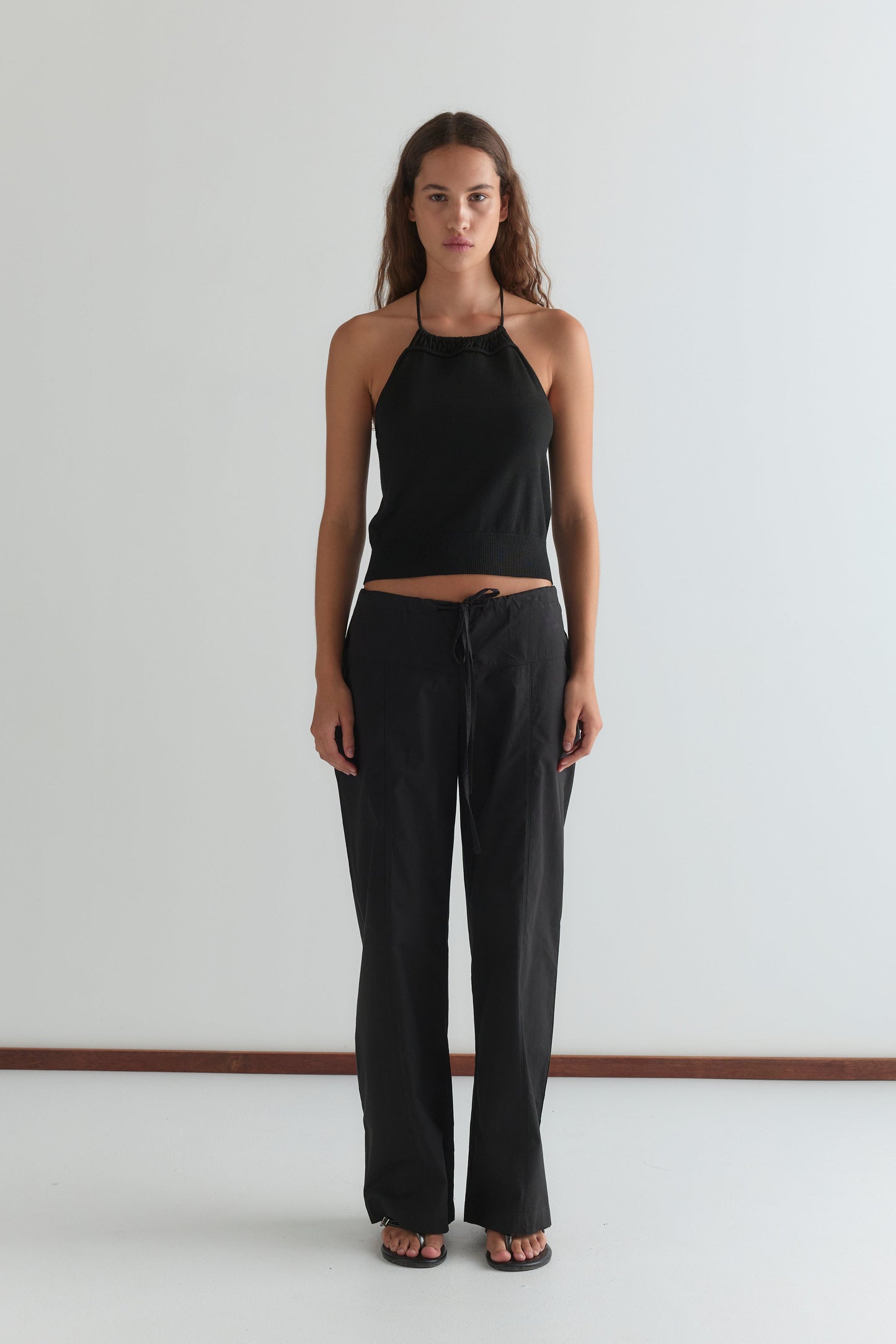 A woman stands against a plain white wall wearing the Deiji Studios Drawcord Knit Top in black, paired with loose black pants and sandals. She has long wavy hair, a neutral expression, and her arms by her sides.