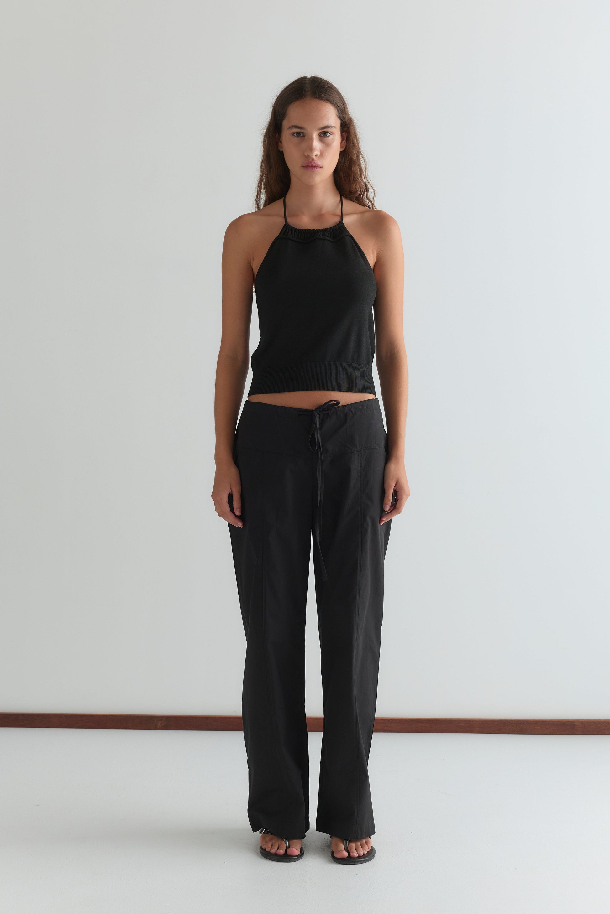 A woman stands against a plain white wall wearing the Deiji Studios Drawcord Knit Top in black, paired with loose black pants and sandals. She has long wavy hair, a neutral expression, and her arms by her sides.
